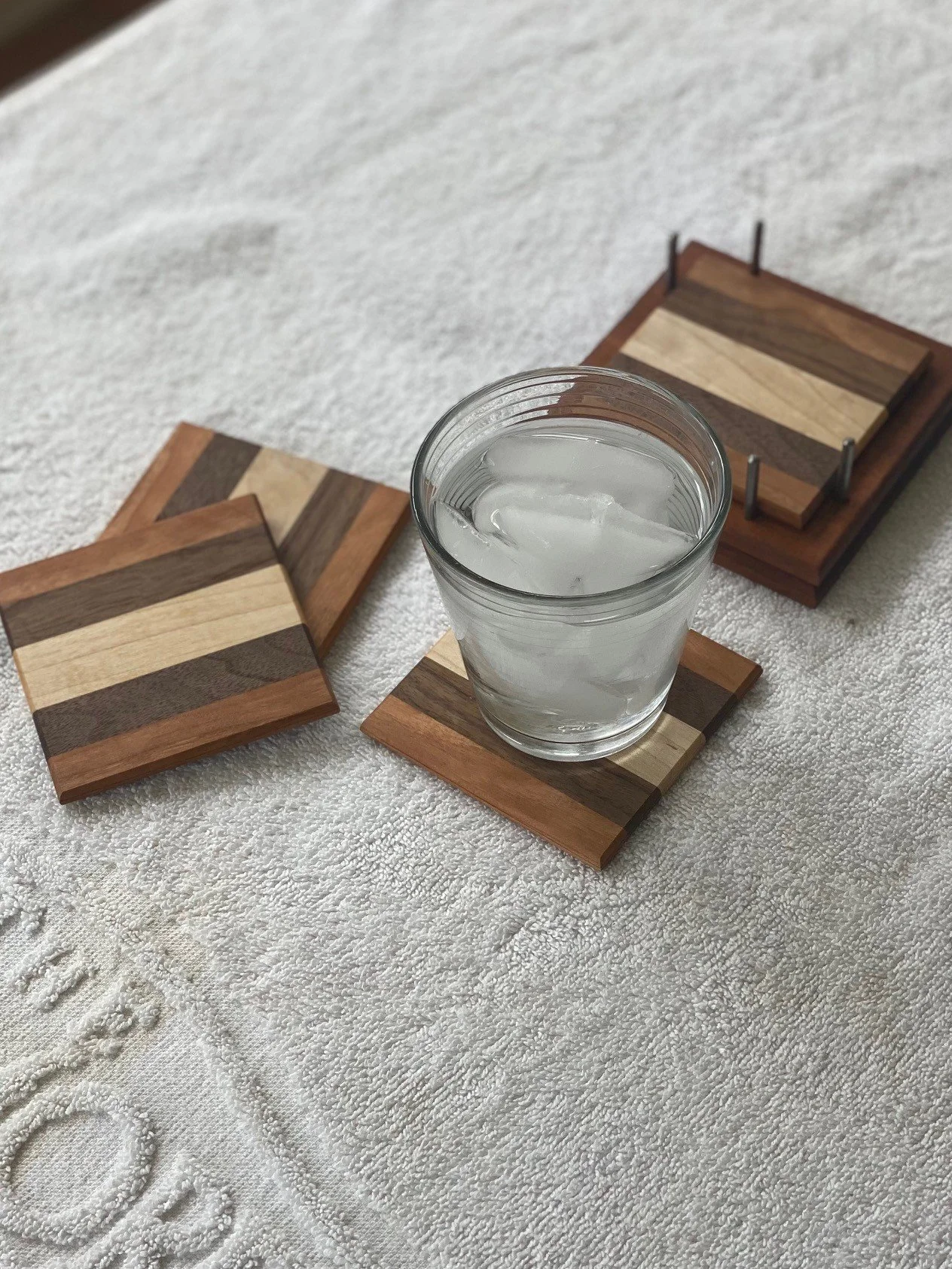 A glass of water with ice cubes on a wooden coaster, surrounded by other wooden coasters on a white textured fabric surface.