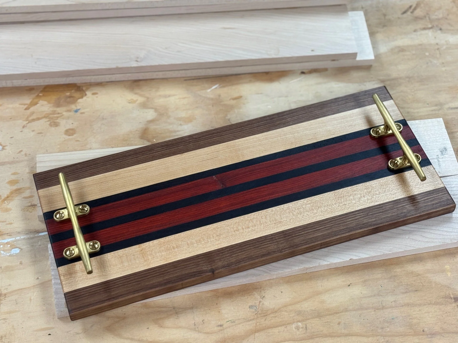 A woodworking project featuring a rectangular piece of wood with a striped pattern of different wood tones, attached with brass hardware, placed on a workbench with other wood pieces in the background.