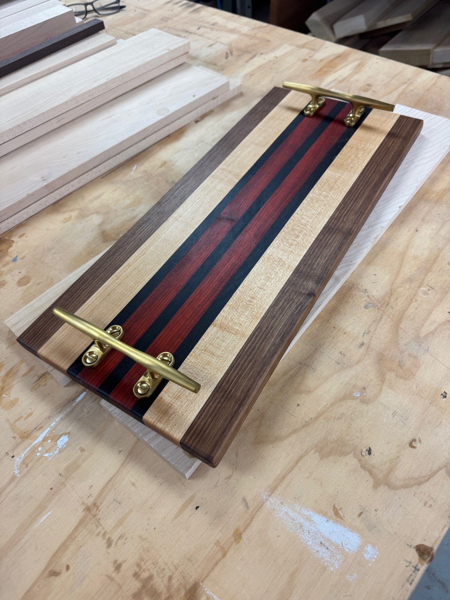 A handcrafted wooden board with a striped pattern of dark and light wood, featuring brass handles on each side, on a woodworking table.