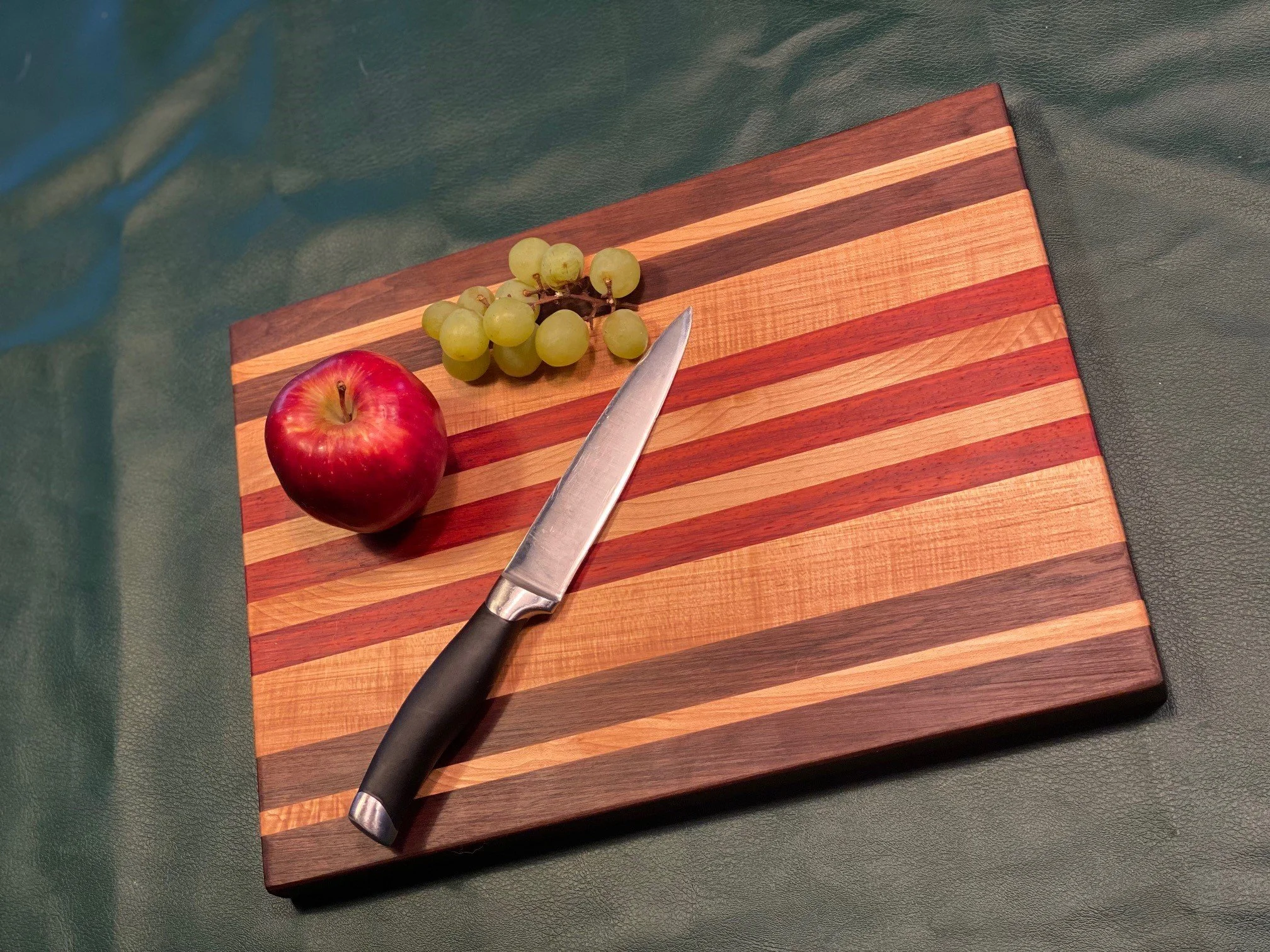A cutting board with colorful wood stripes, a bunch of green grapes, a red apple, and a knife on top, placed on a dark green surface.