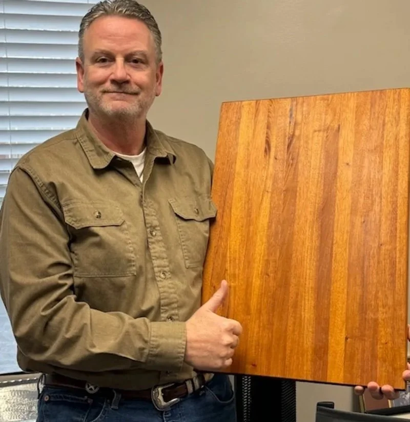 A man with gray hair and a beard, wearing a tan button-up shirt, giving a thumbs up while holding a large wooden panel with a polished finish in a room with beige walls and a window with blinds.