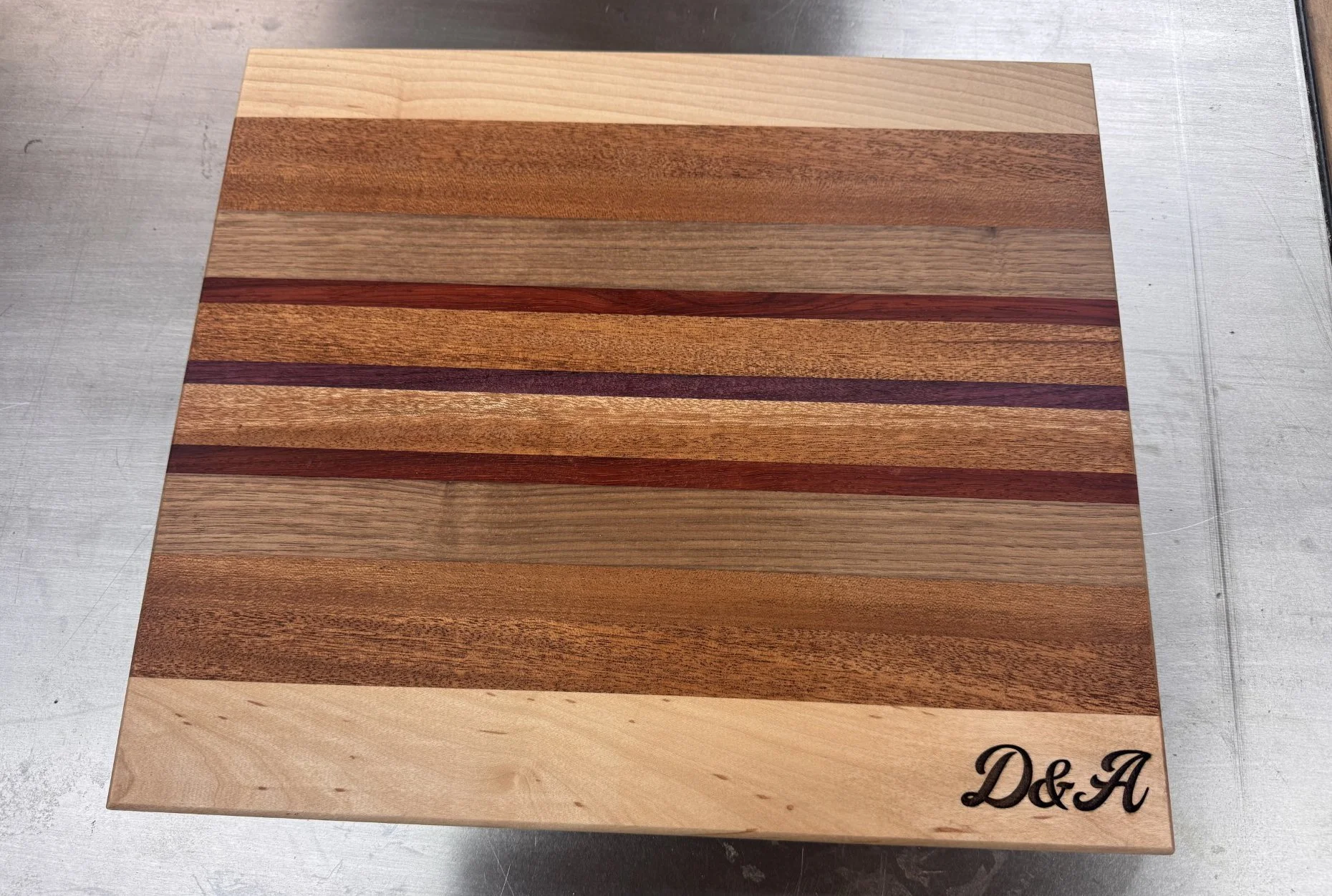 A wooden cutting board with multiple horizontal strips of different types and shades of wood. The bottom right corner of the board has the initials 'D&A' in black script.