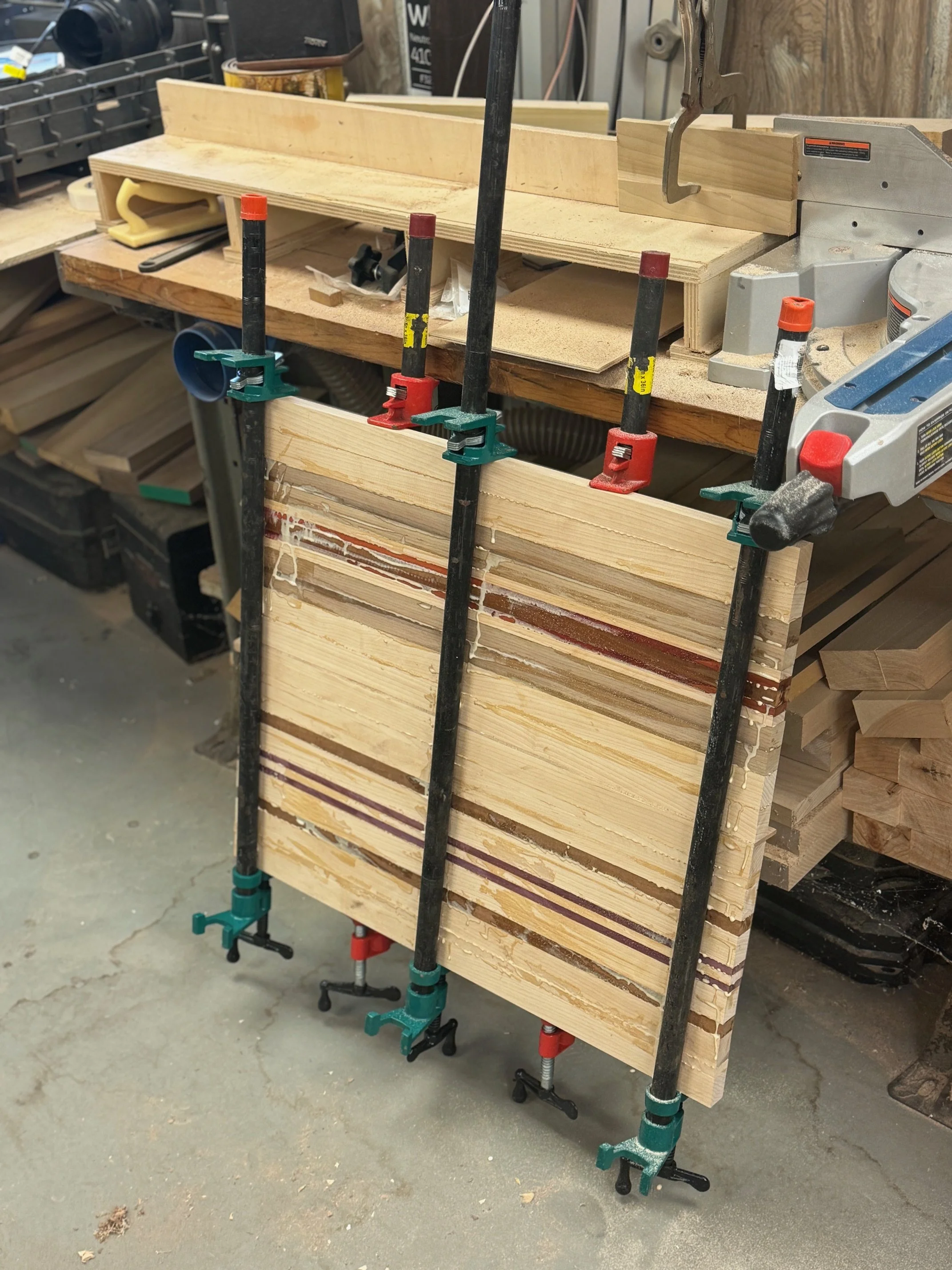 A woodworking workbench with clamps securing a stack of wooden boards and panels, in a workshop setting.