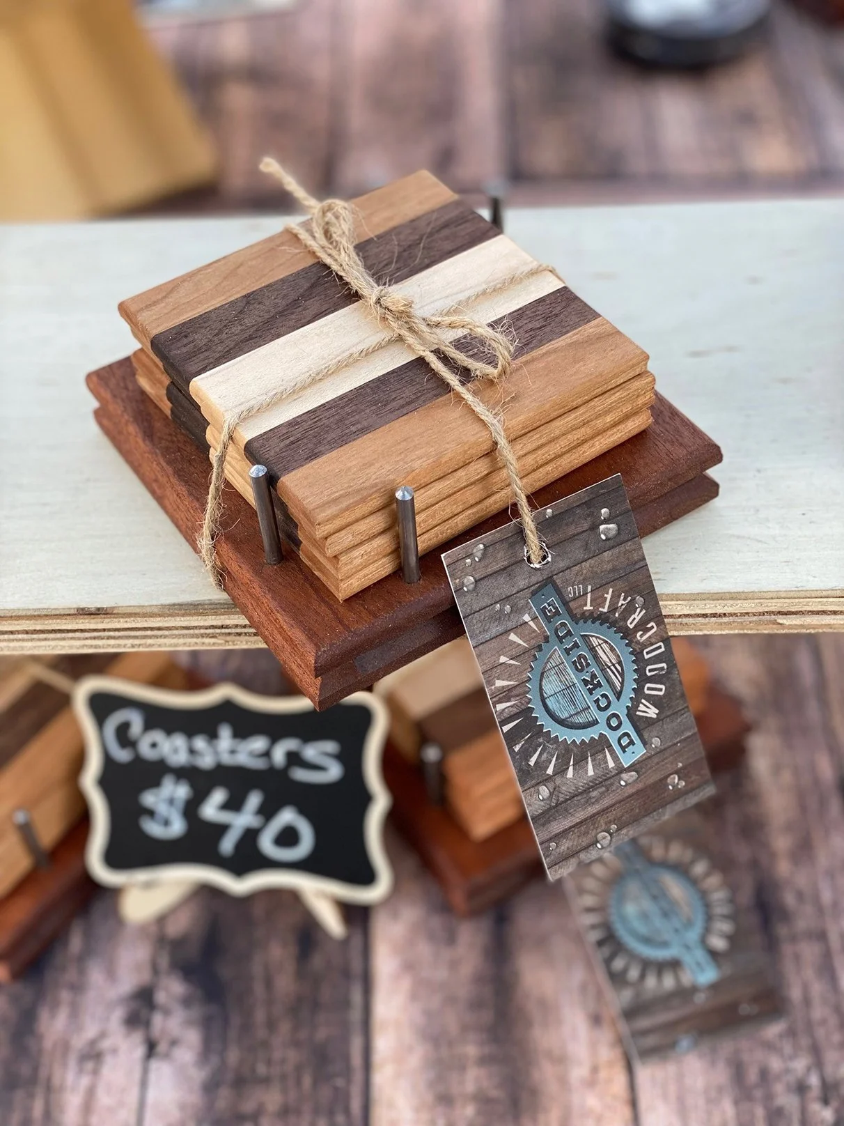 Stack of small wooden boards tied with string on a display stand, with a tag that reads 'DISCOUNTED WOODEN COASTERS' and a handwritten chalk sign indicating $40 for coasters.
