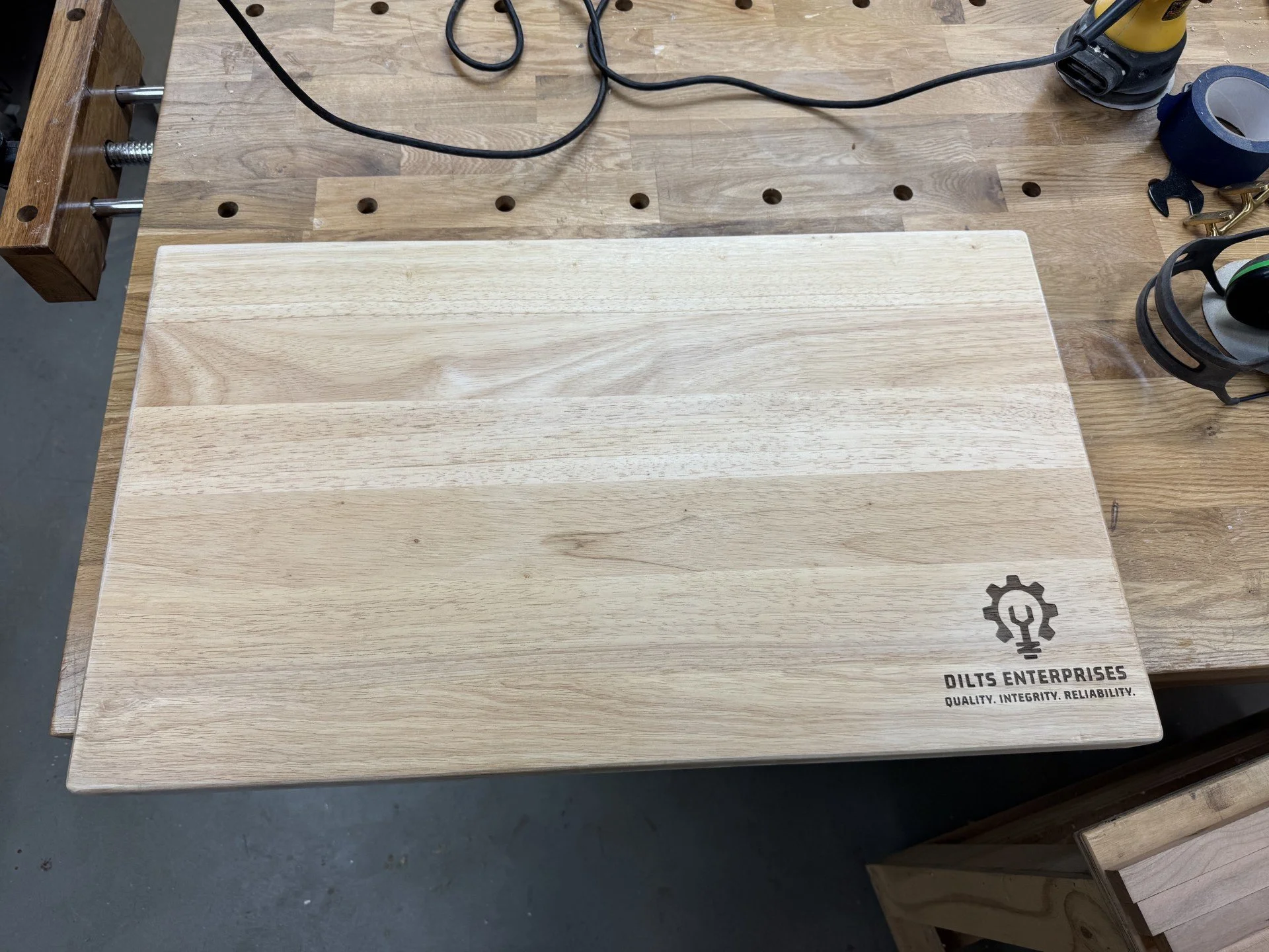 A wooden workbench with evenly spaced holes, holding a light-colored wooden board with the logo and text 'DILTS ENTERPRISES' and 'QUALITY, INTEGRITY, RELIABILITY'. Various tools, black cords, electrical equipment, and rolls of tape are visible on the