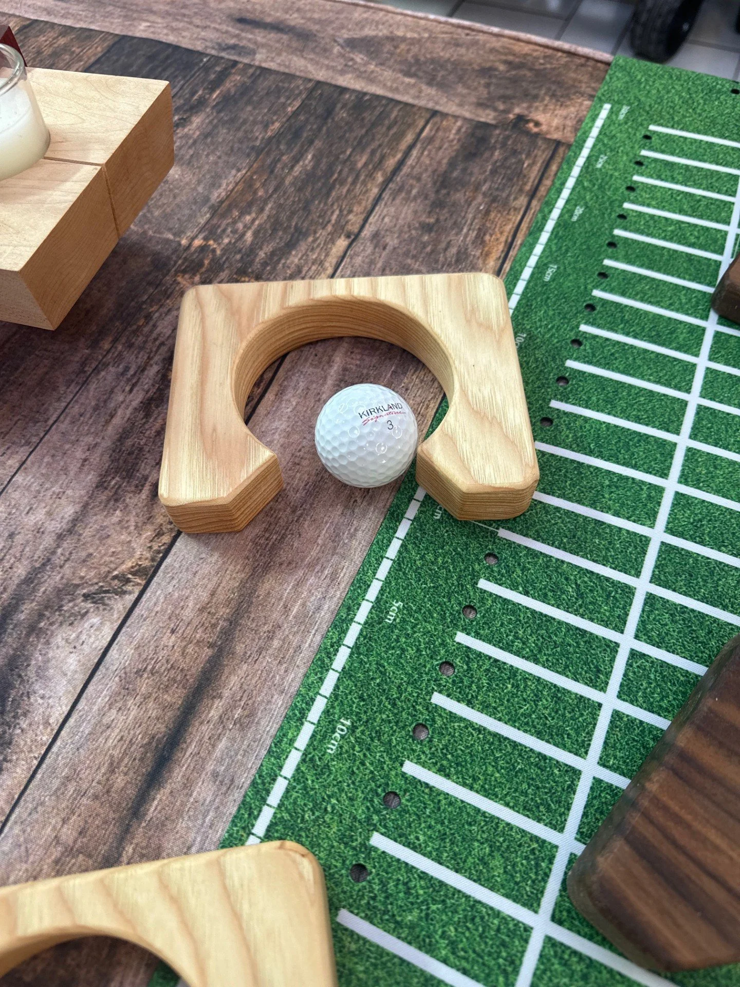 A golf ball resting on a green practice mat, with wooden blocks and a small wooden chute.