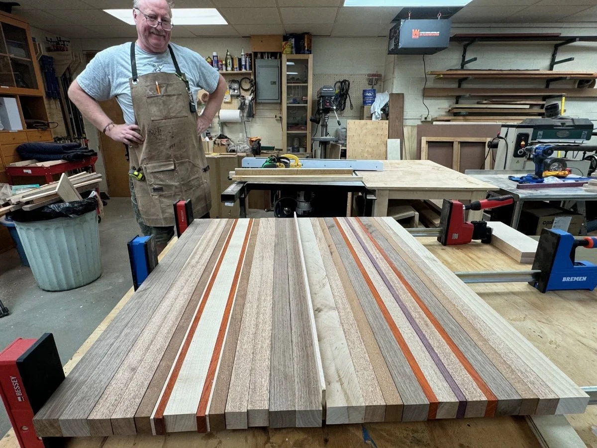 A man wearing a gray t-shirt and an apron stands in a woodworking workshop, smiling. In the foreground, there is a large wooden workpiece with striped patterns clamped to a workbench. The background shows woodworking tools and equipment, shelves with supplies, and various pieces of wood and machinery.
