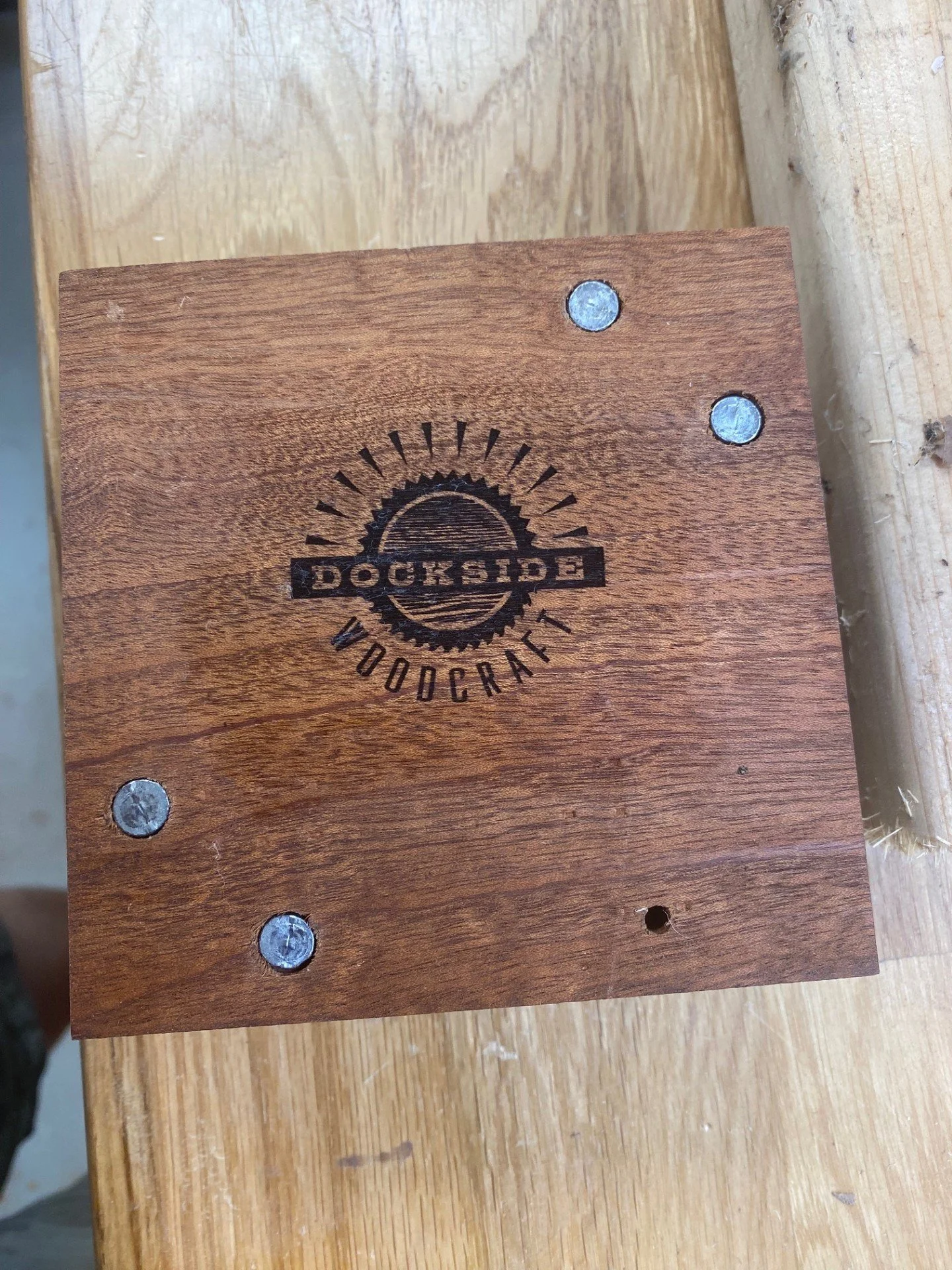 Close-up of a wooden piece with a logo that reads 'Dockside Woodcraft', corner screws, and a hole at the bottom right corner.