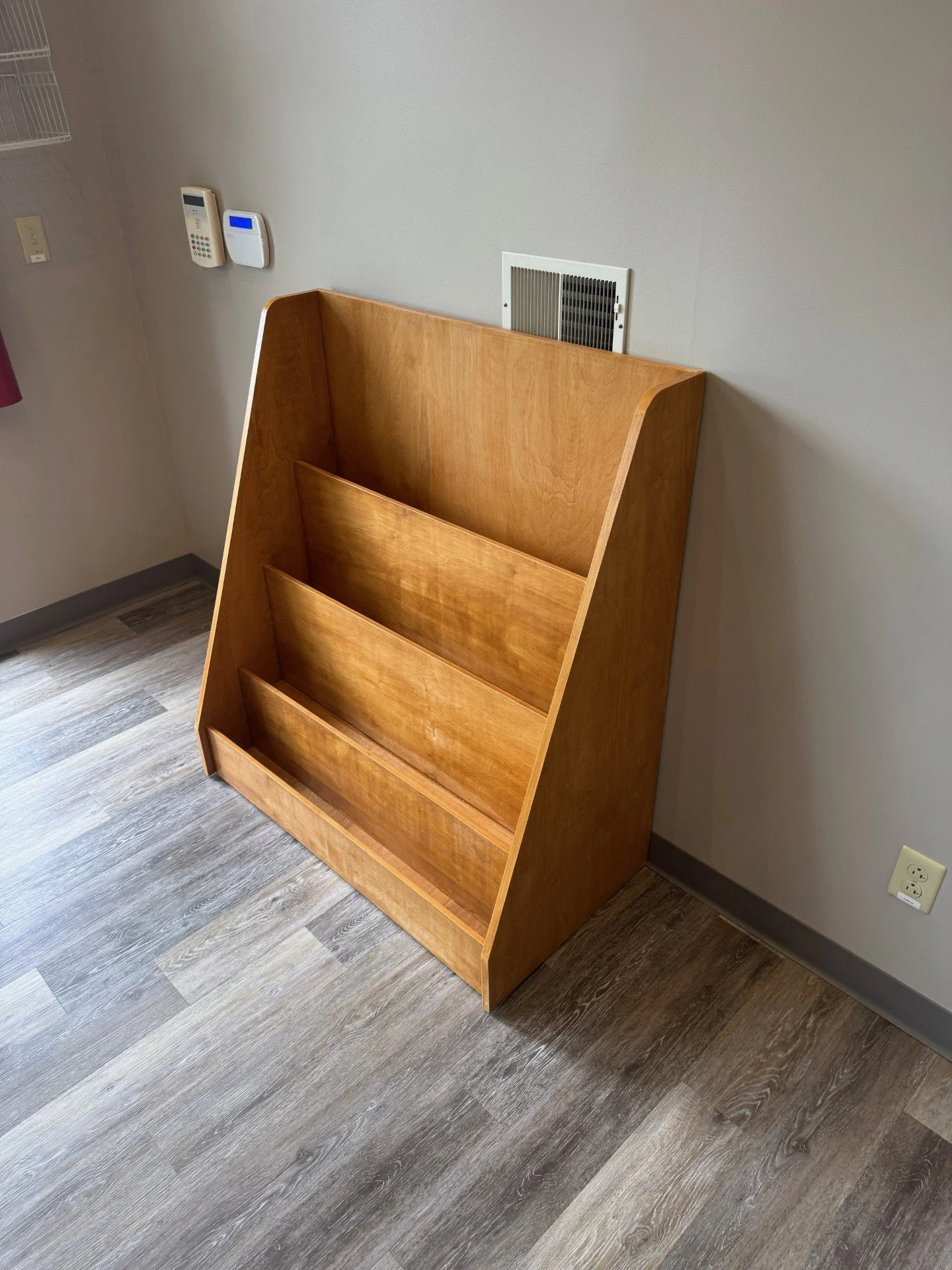 Empty wooden bookshelf standing next to a wall with a vent and electrical outlets in a room with wood-look flooring.