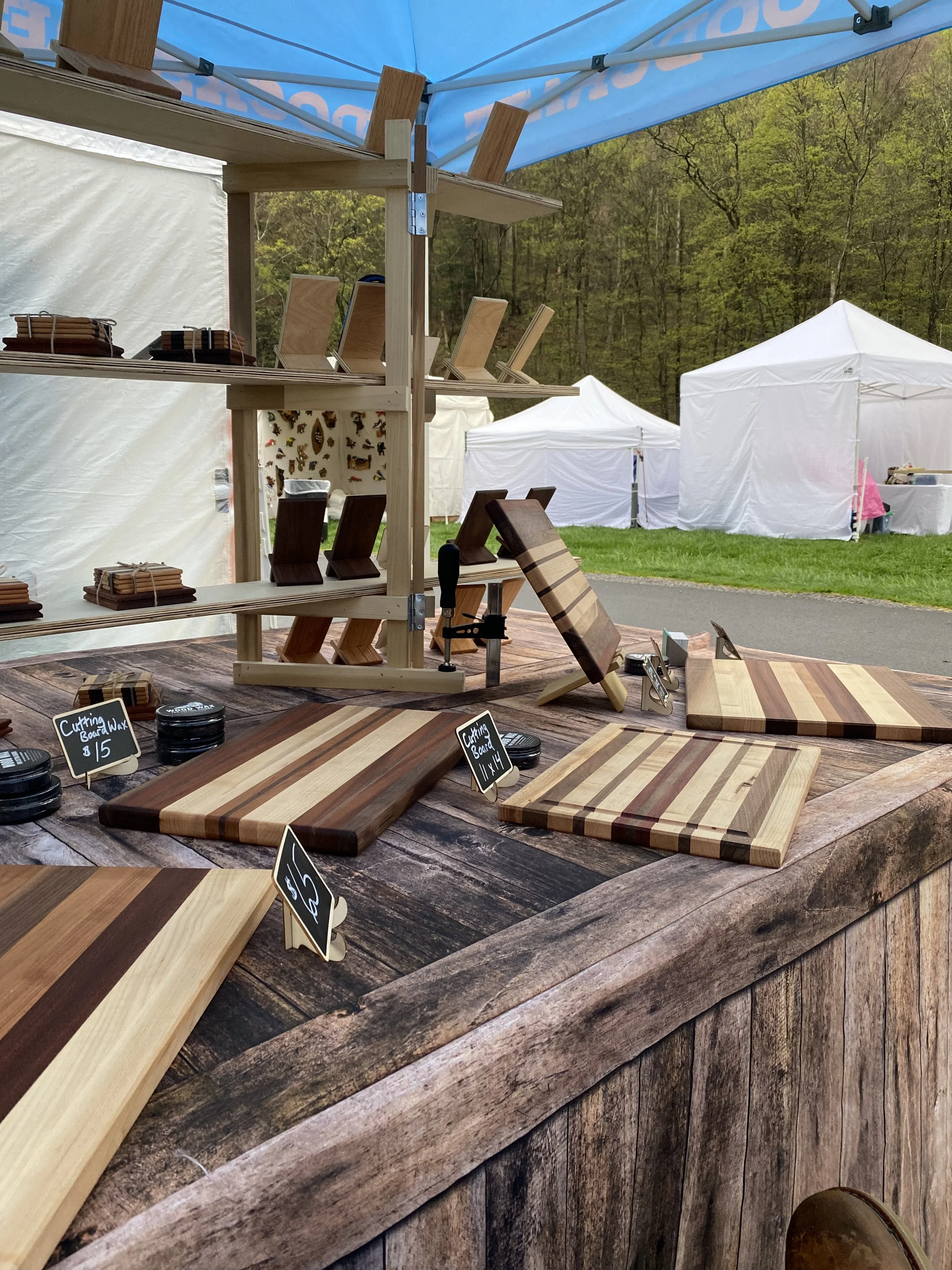Display of various wooden cutting boards on a rustic wooden table at an outdoor market, with tents and trees in the background.