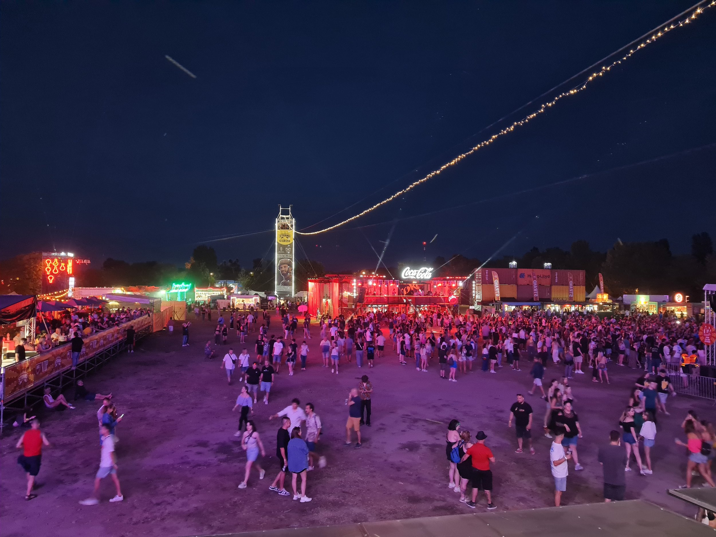 Crowd of people at a large outdoor event during nighttime, with colorful lights, tents, and stalls. Two illuminated Slider zipline tracks stretch across the sky above the event area, creating a spectacular aerial attraction. A clear night sky with vi
