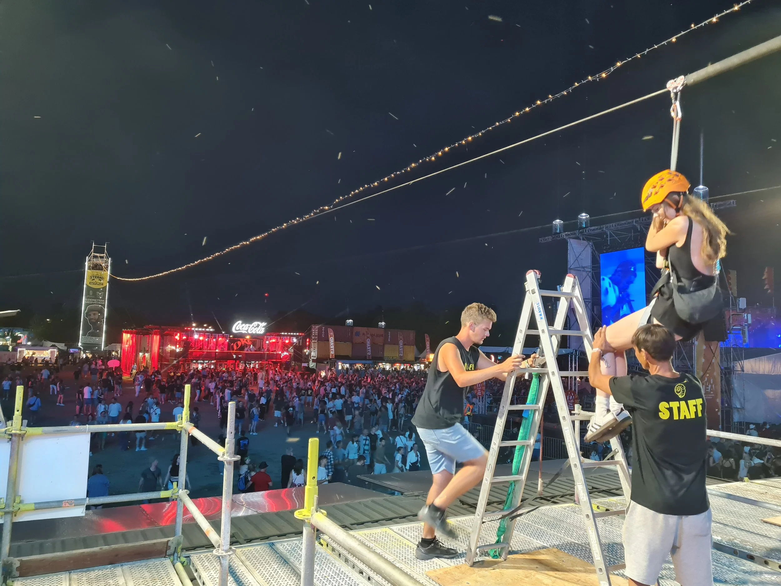 A large outdoor nighttime event with a crowd, vibrant lights, a stage area, and crew members working on scaffolding during setup. Two Slider mobile zipline tracks extend above the venue, creating a spectacular aerial experience and a unique visual el