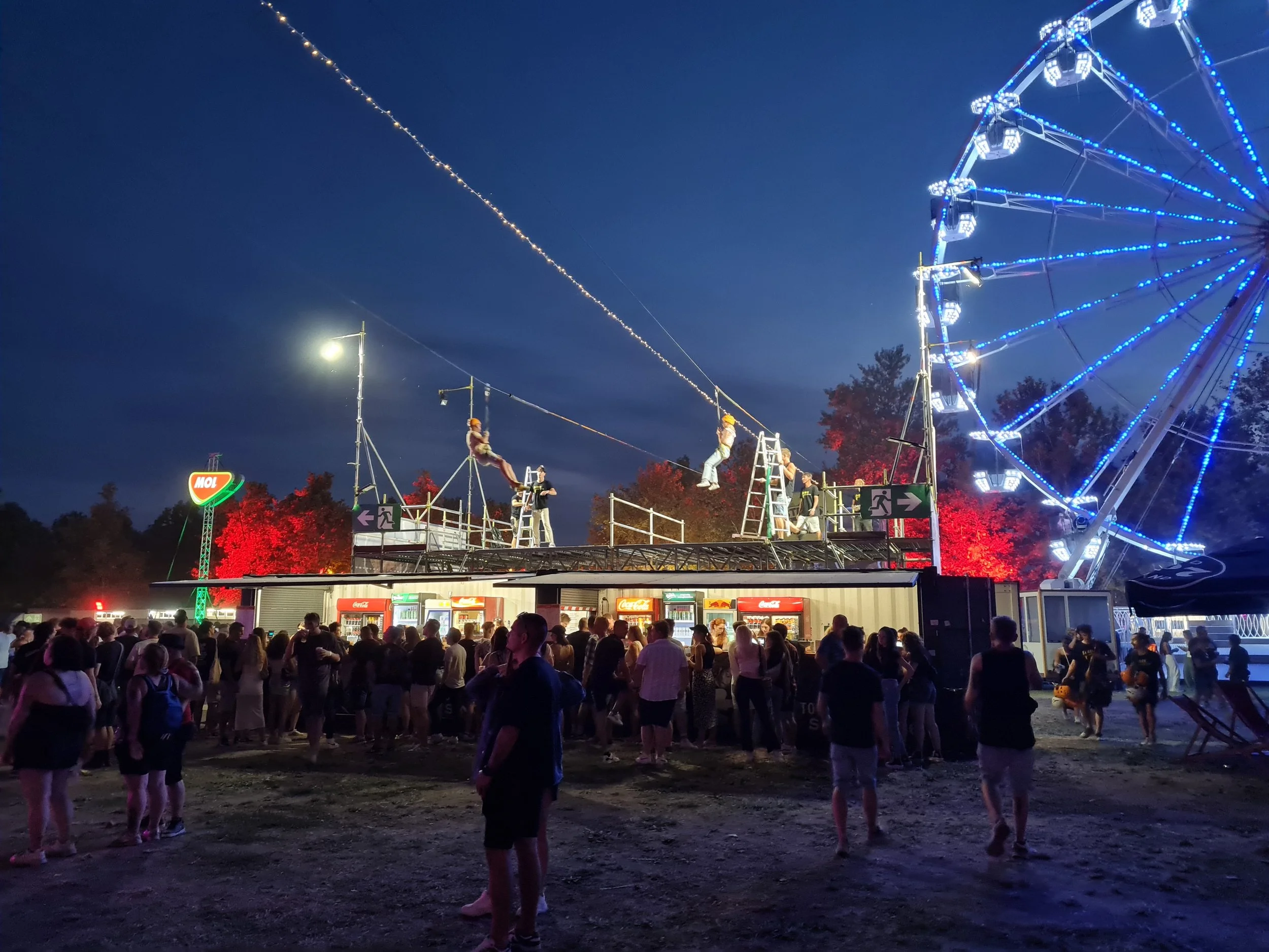 A vibrant nighttime festival scene with a large crowd, illuminated fairground rides, colorful lights, and an energetic atmosphere. Two Slider mobile zipline tracks stretch above the event area, creating a unique aerial attraction that complements the