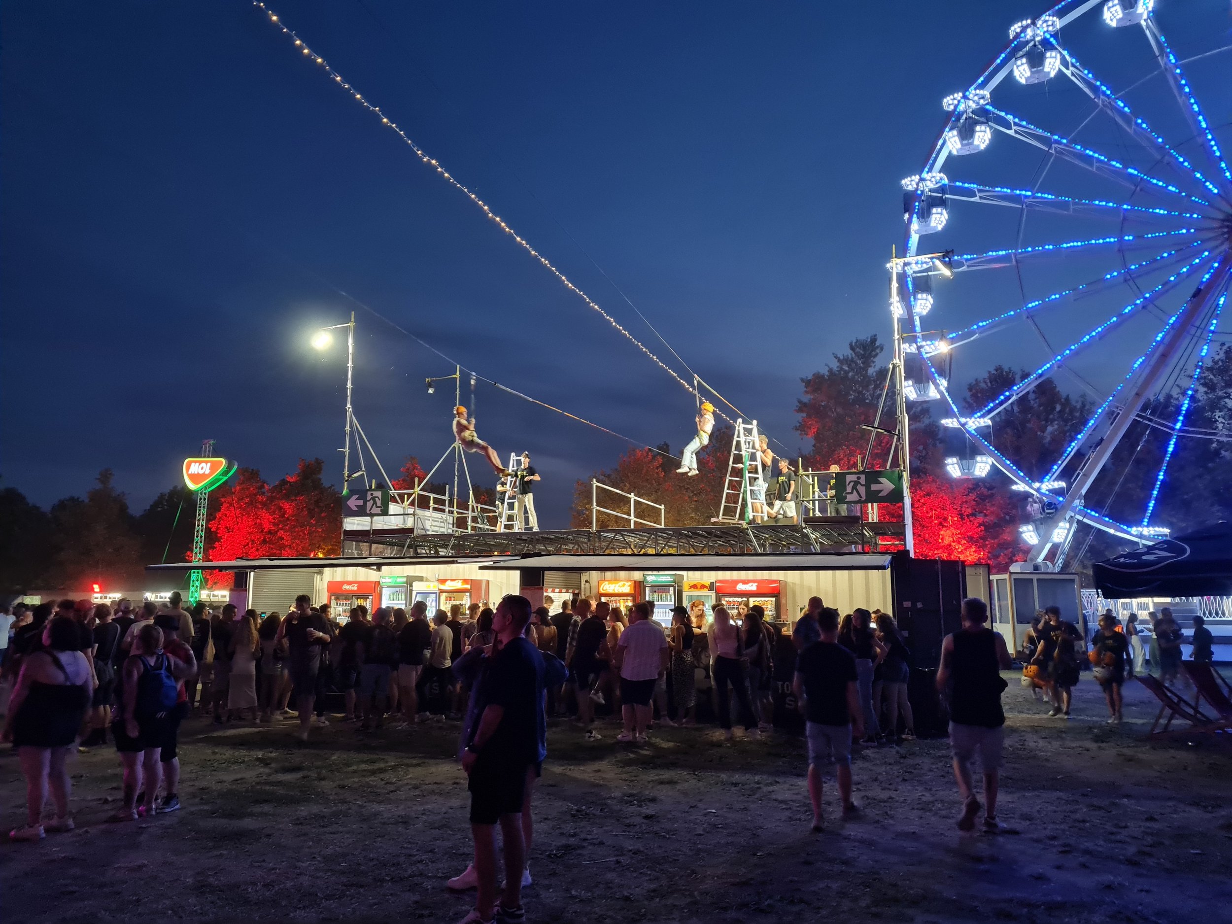 A vibrant nighttime festival scene with a large crowd, illuminated fairground rides, colorful lights, and an energetic atmosphere. Two Slider mobile zipline tracks stretch above the event area, creating a unique aerial attraction that complements the