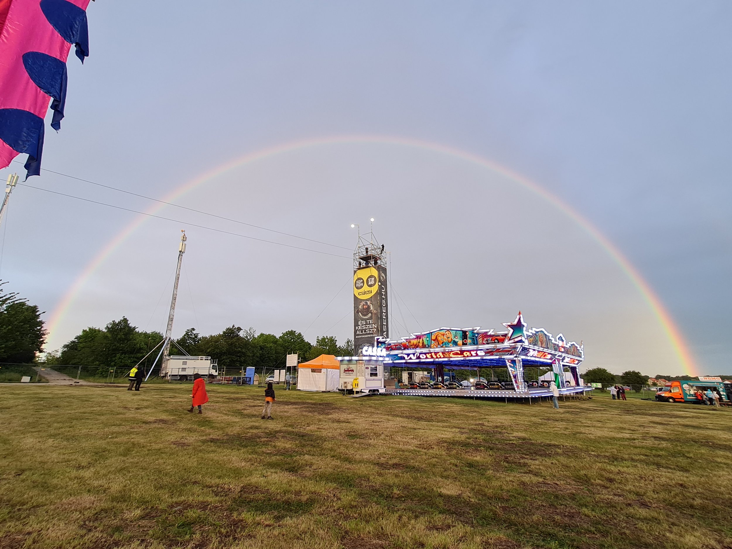 A colorful fairground scene with a rainbow stretching across the sky, visitors exploring the event area, and various attractions surrounded by tents and vehicles. Two Slider mobile zipline tracks stand out above the venue, adding a spectacular aerial