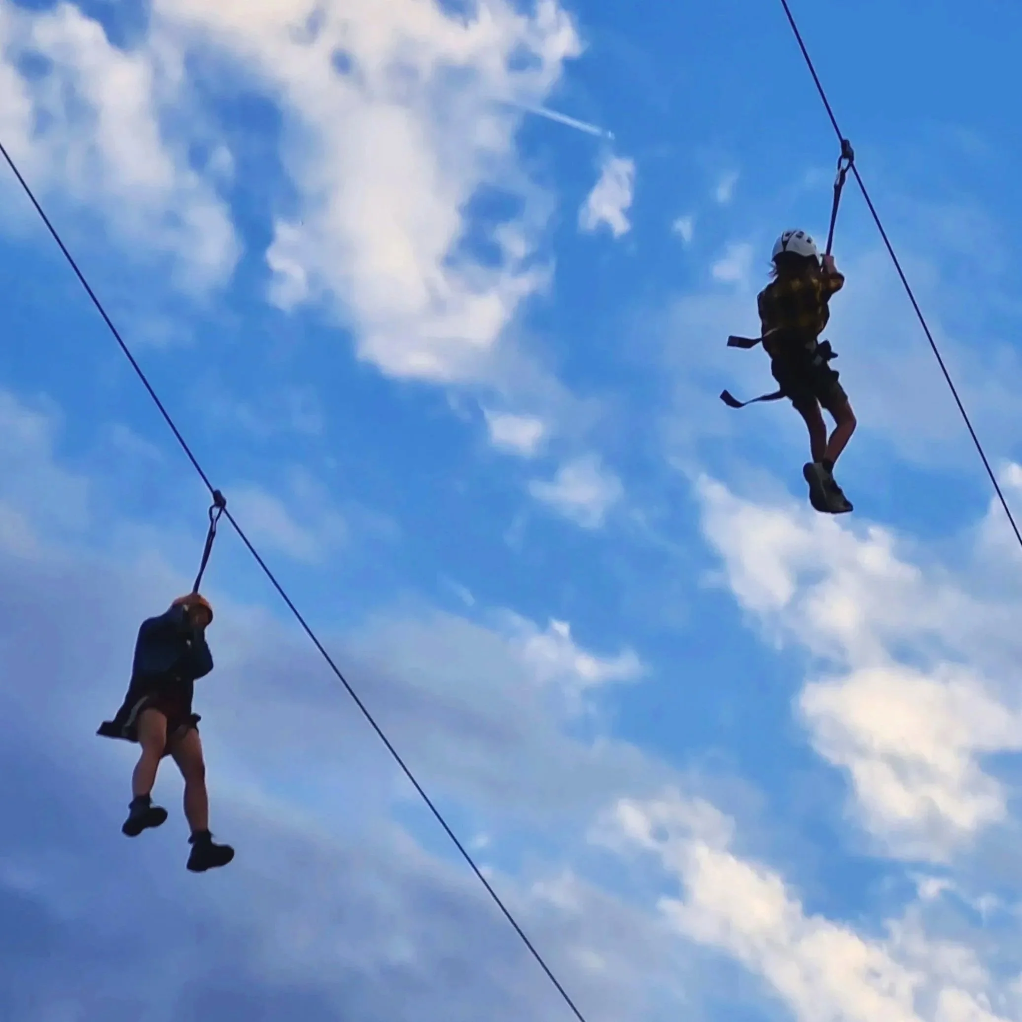 Two individuals, wearing helmets, are walking on a high ropes course against a blue sky with clouds. They are attached to safety cables and appear to be navigating the suspended ropes or platforms.