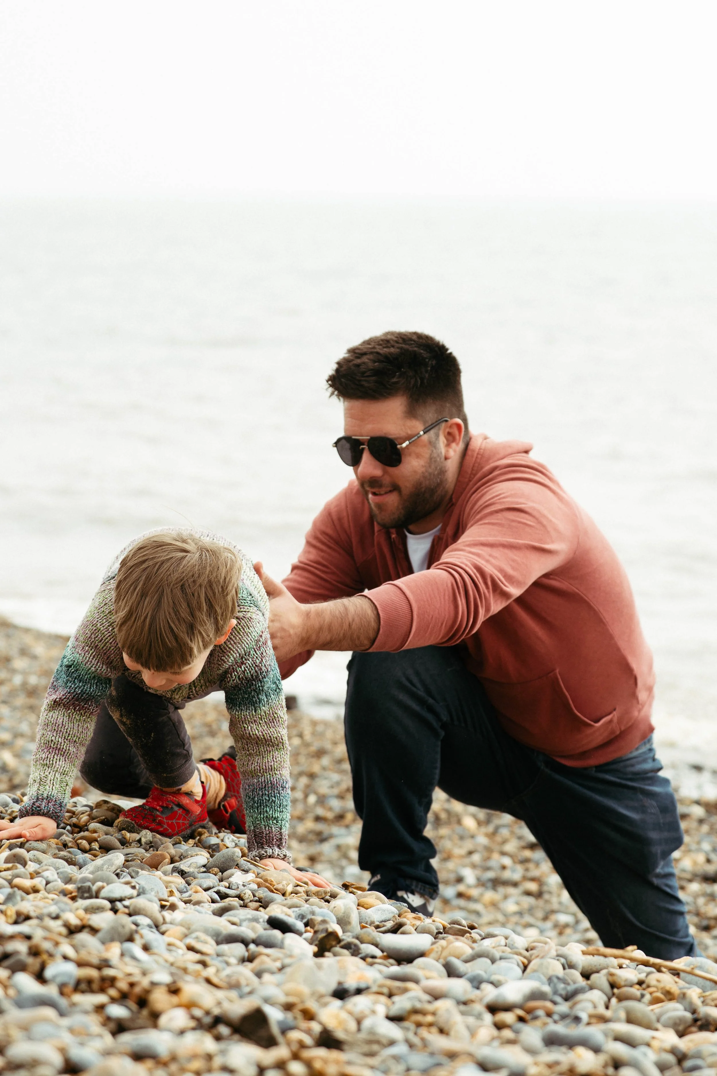 A man wearing sunglasses and a reddish jacket crouches on a pebble beach, helping a young boy in a colorful sweater and red shoes pick up stones, with the ocean visible in the background.