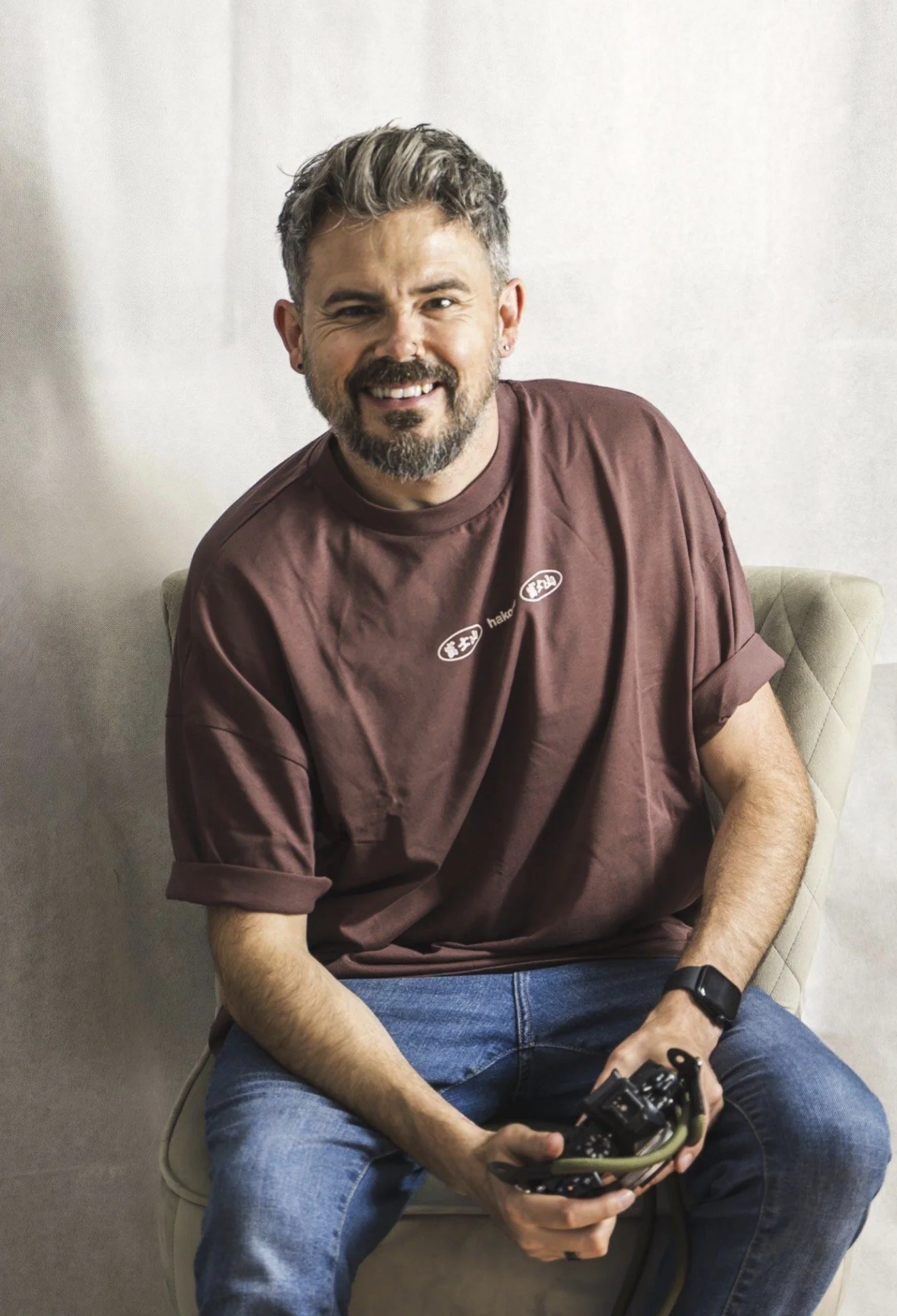 A smiling man with a beard and a mustache, wearing a brown t-shirt and jeans, sitting on a chair against a plain wall, holding a camera in his hands.