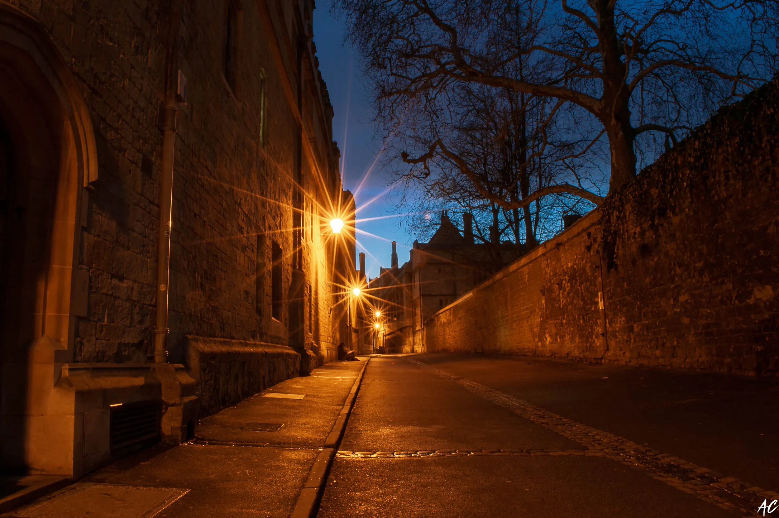 A quiet street at night illuminated by streetlights, with bare trees and old stone buildings on either side.