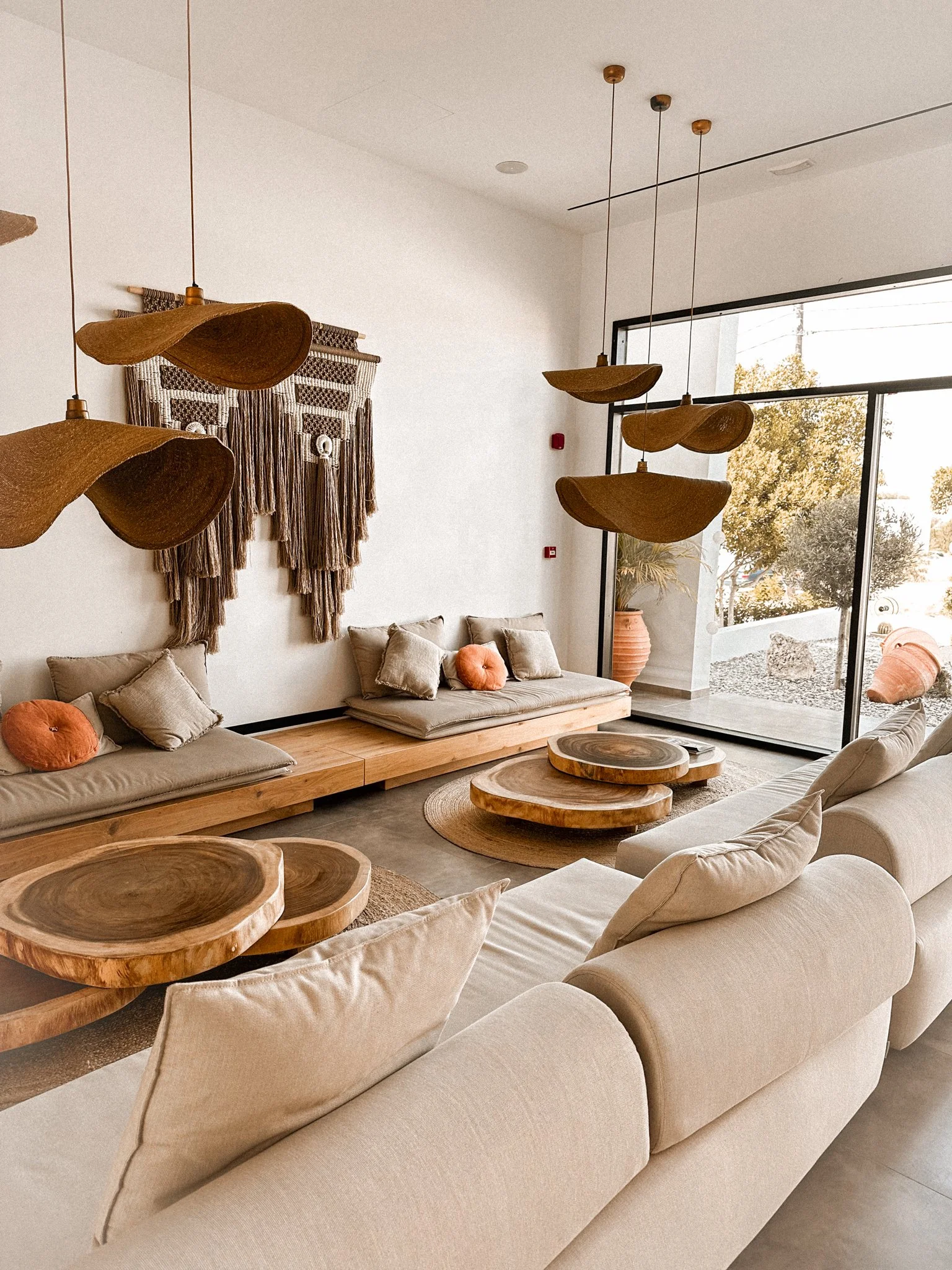 Living room with beige cushioned sofas, wooden coffee tables, hanging wooden light fixtures, and a wall hanging with dark brown and beige tassels. Large glass window shows trees outside.