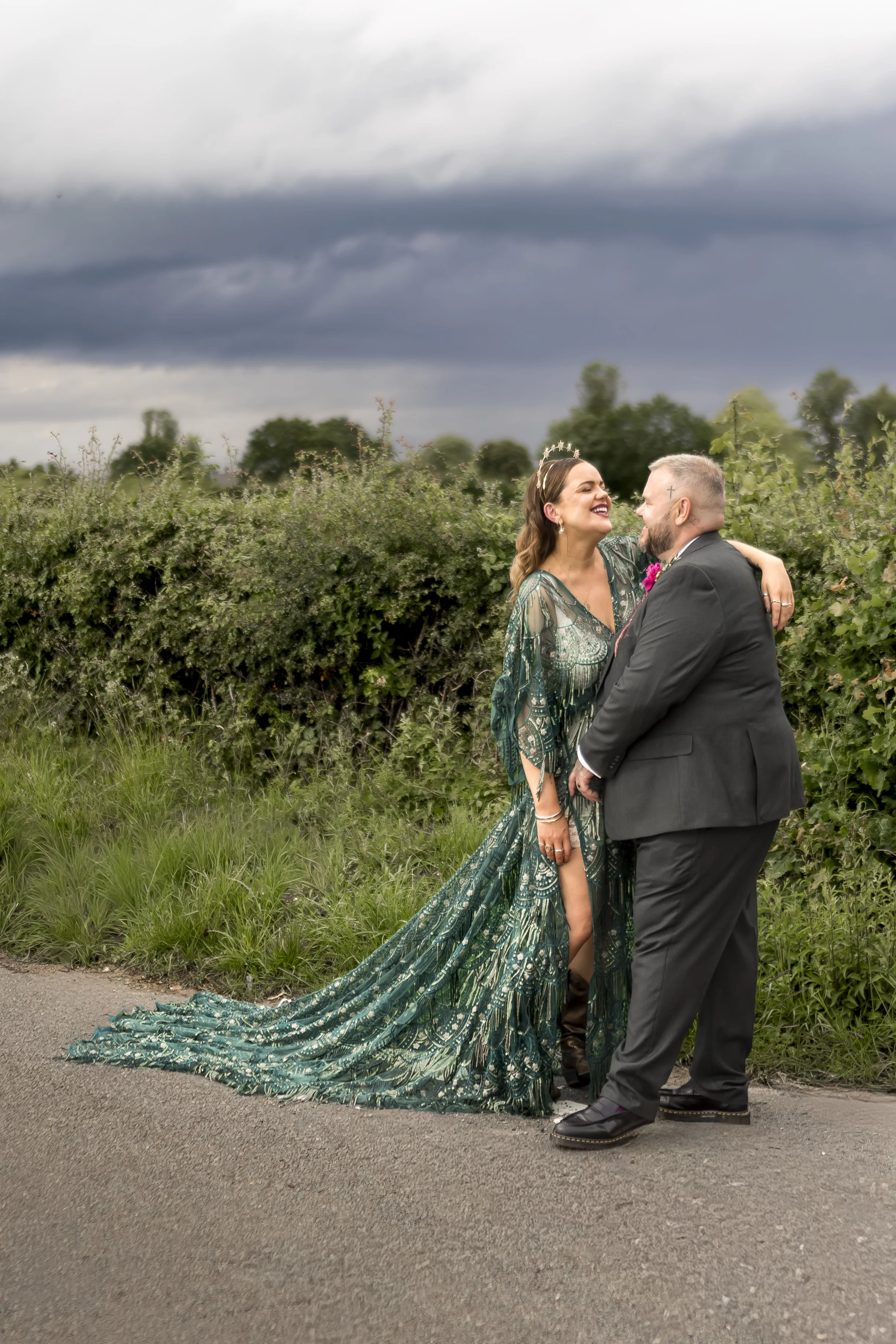 A man and woman in wedding attire stand close together outdoors, smiling and looking at each other. The woman wears a long, flowing green and gold patterned dress with a plaid train, and the man wears a dark suit with a pink boutonniere. The backgrou