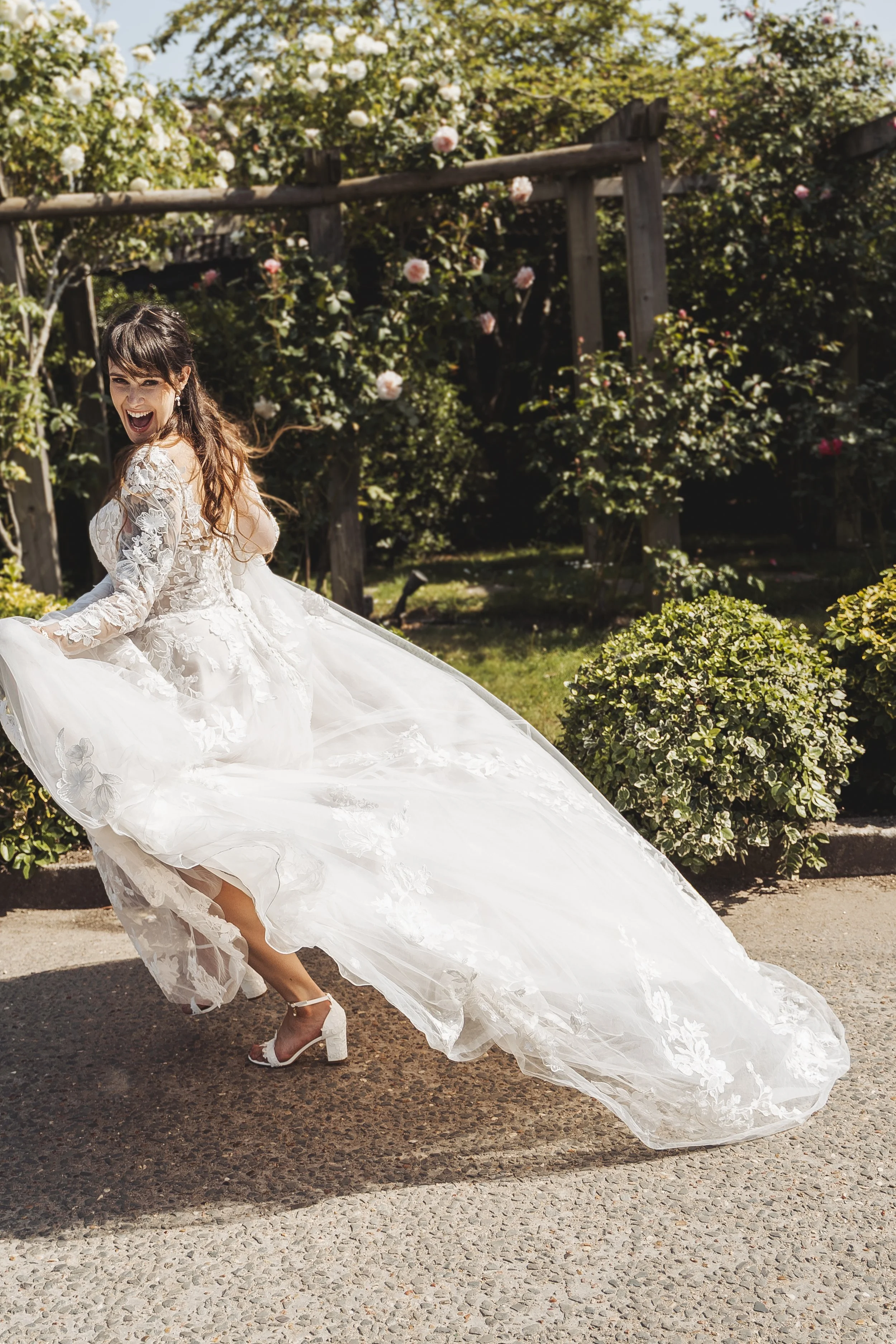 A woman in a white wedding dress with lace details and heels is smiling and twirling outdoors in a garden with flowering bushes and trees.