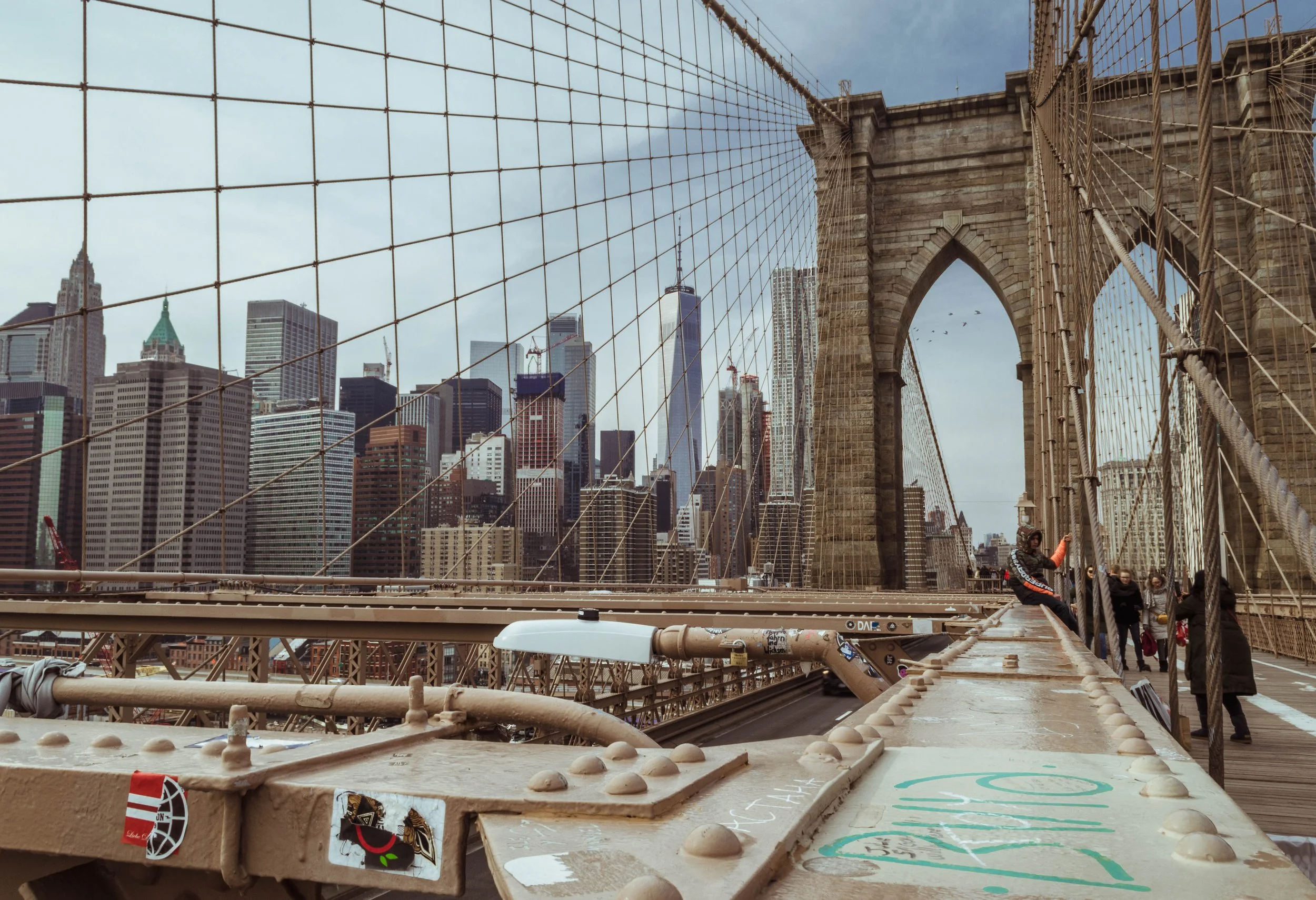 View of Brooklyn Bridge with the Manhattan skyline in the background, including tall buildings and skyscrapers. People are walking on the bridge, and the bridge's stone arch and suspension cables are visible.