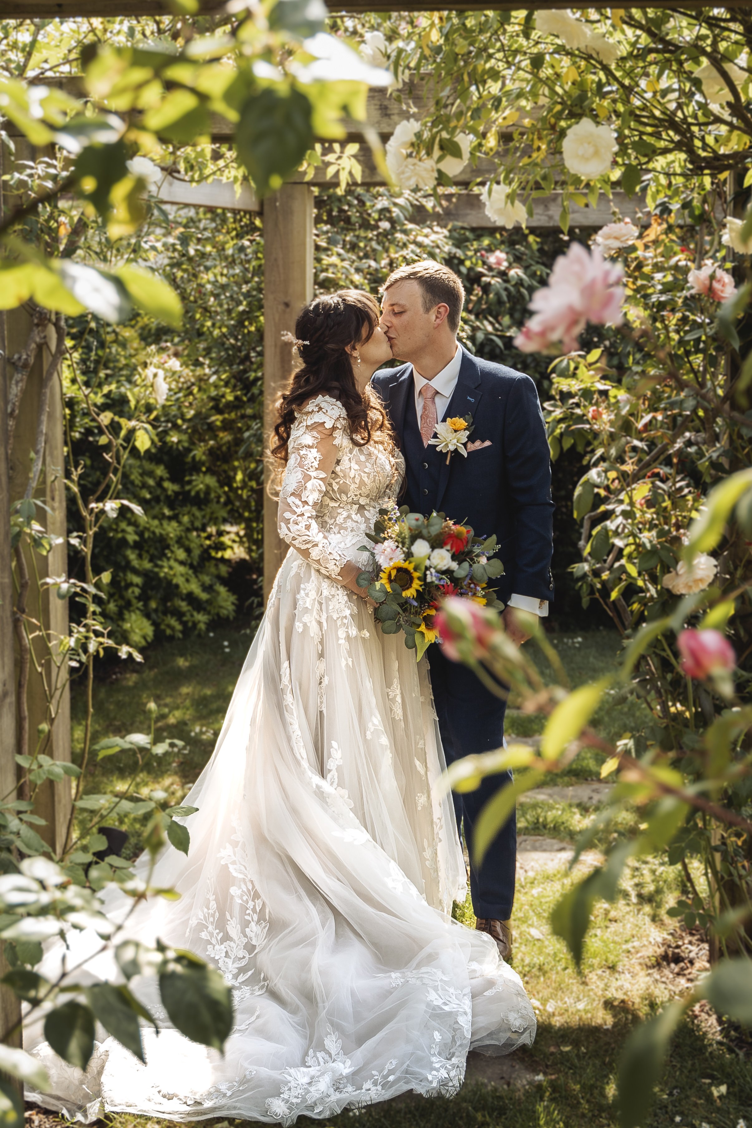 A bride and groom share a kiss in a garden surrounded by blooming flowers, with the bride in a lace wedding gown and the groom in a navy suit.