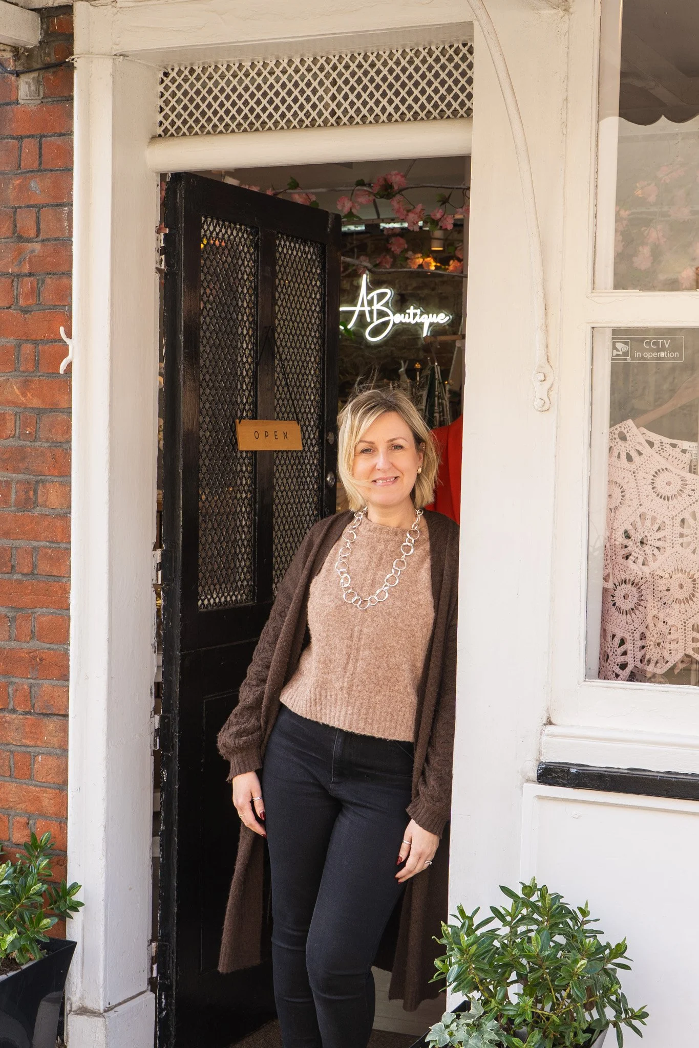 A woman with blonde hair wearing a brown cardigan, beige sweater, and black pants, standing outside  Aboutique store with a black door, floral decorations inside, and potted plants outside.