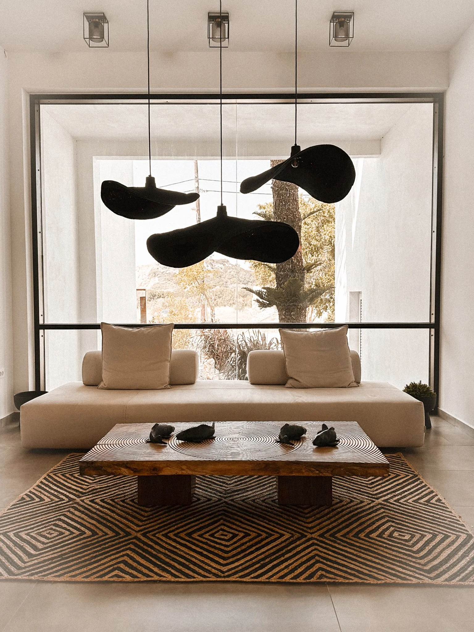 Living room with beige sofa, four dark decorative stones on a striped black and brown rug, large glass window with trees outside, hanging black pendant lights, and a wooden table.