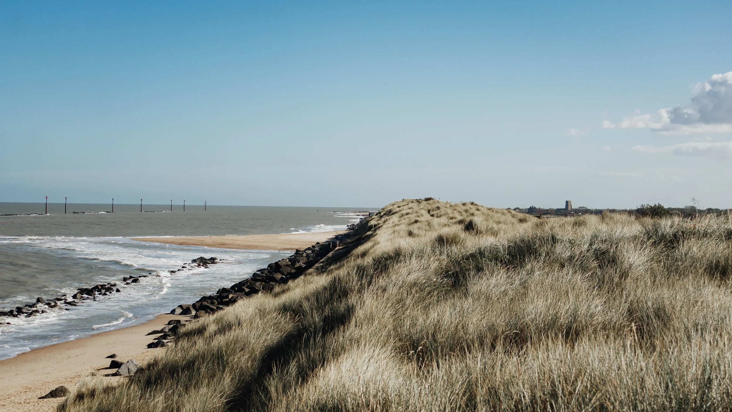 A coastal landscape with sandy dunes covered in grass, rocky shoreline, ocean waves, and a partly cloudy sky.