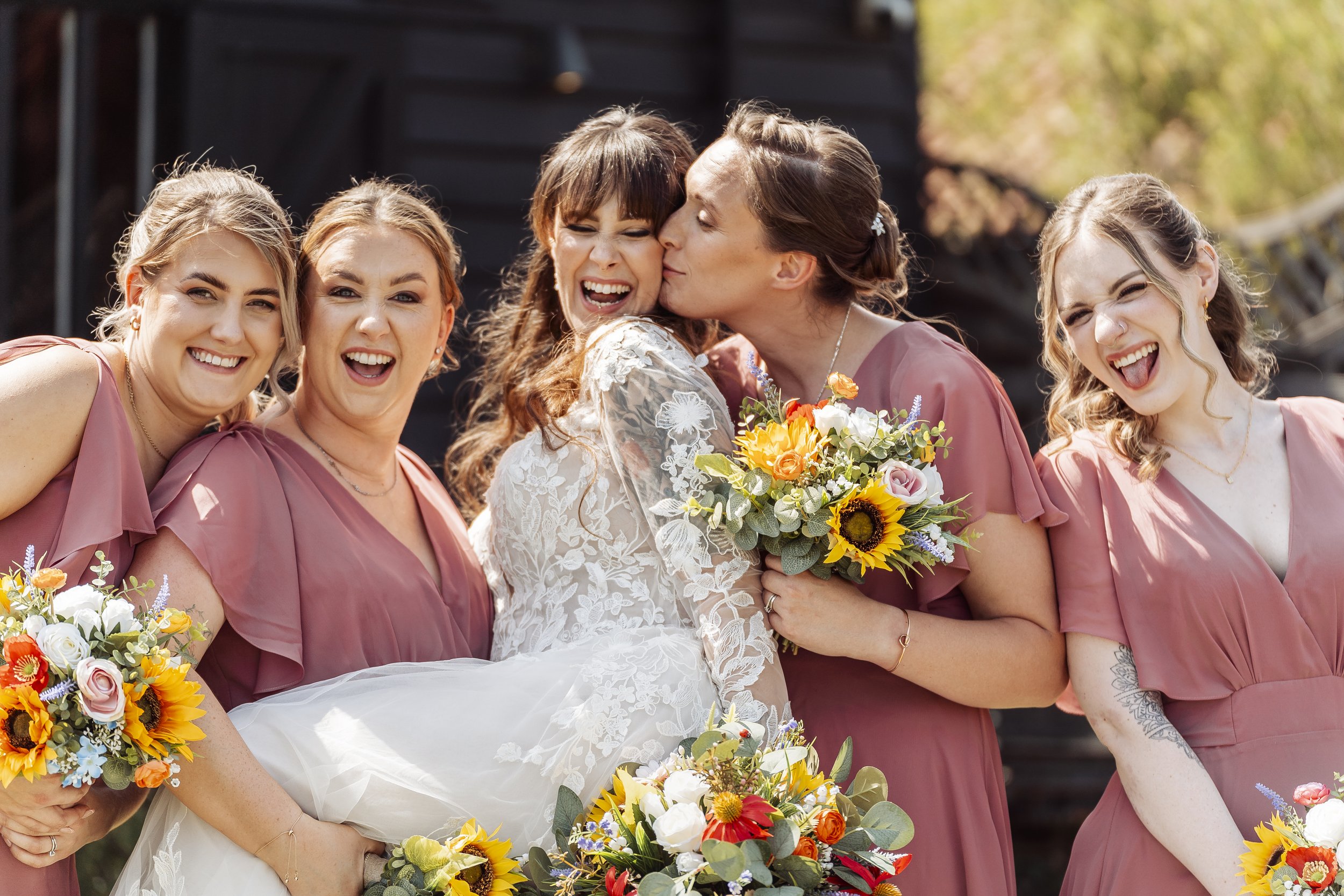 A group of women, including a bride in a white lace dress, celebrating at a wedding outdoors, holding bouquets of flowers with sunflowers and other colorful blooms.