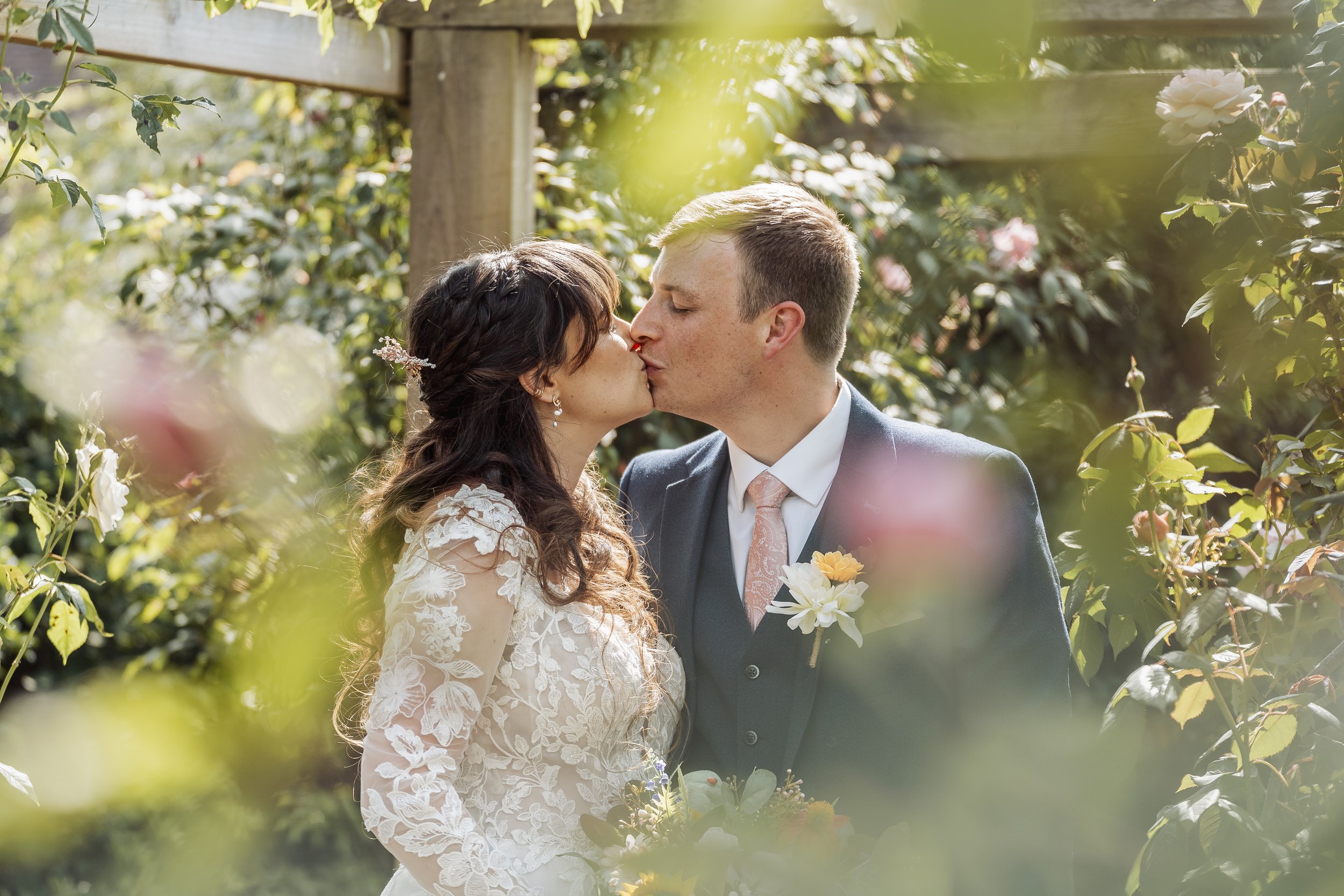 A bride and groom share a kiss in a garden surrounded by greenery and flowers, with sunlight filtering through the leaves.