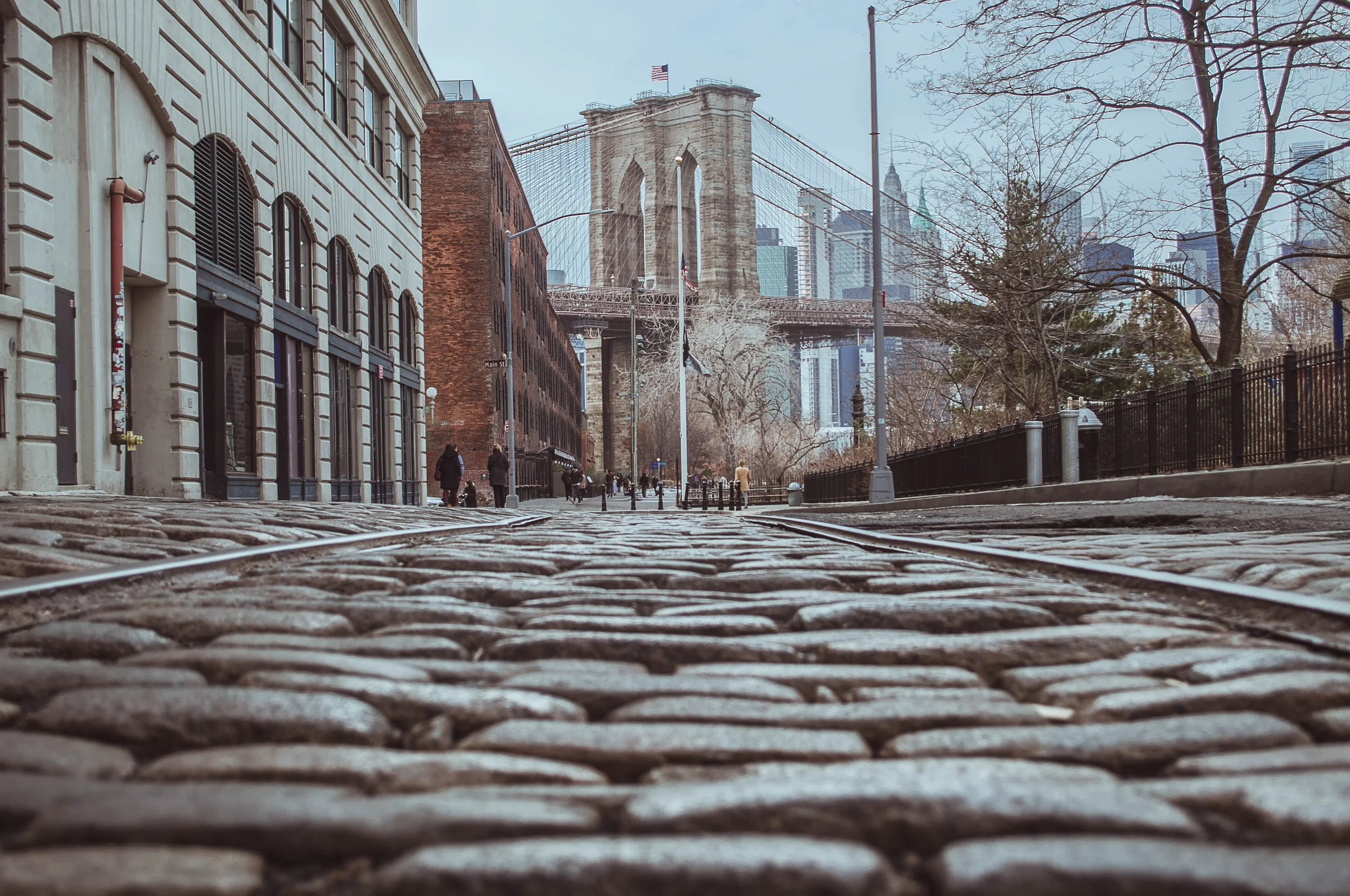 View of Brooklyn Bridge in New York City with cobblestone street foreground, historic buildings, and city skyline in the background.
