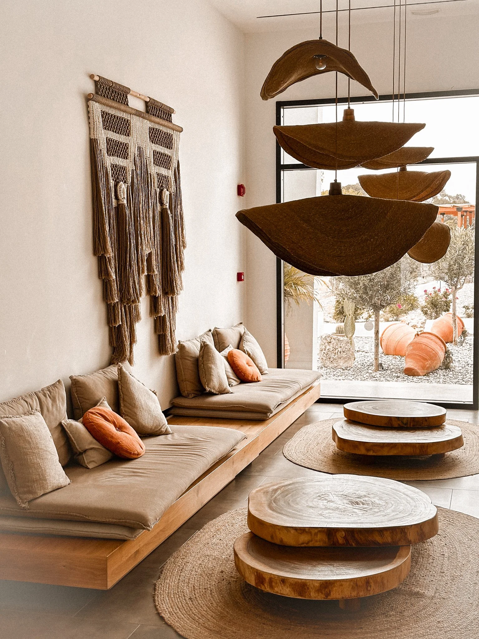 A living room with beige cushioned seating, orange pillows, a matching beige throw, and a round woven rug. Wooden tables with natural wood finishes and rounded edges are in the foreground. A large window with a view of outdoor pottery and desert plan