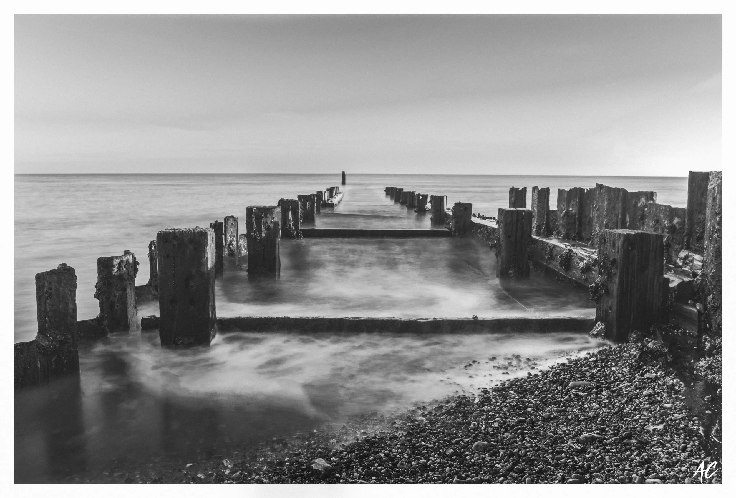Black and white photograph of a seascape with a wooden breakwater extending into the water, ending in a solitary post, and a rocky shoreline in the foreground.