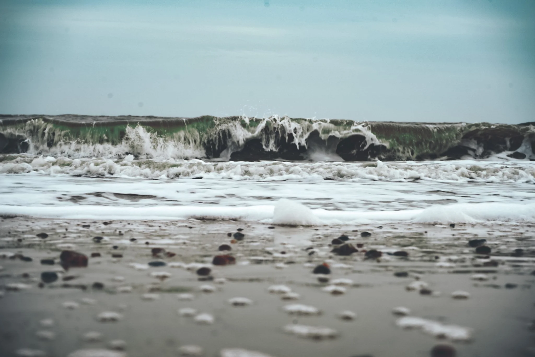 Ocean waves crashing onto the sandy shoreline with small rocks and pebbles, under an overcast sky.