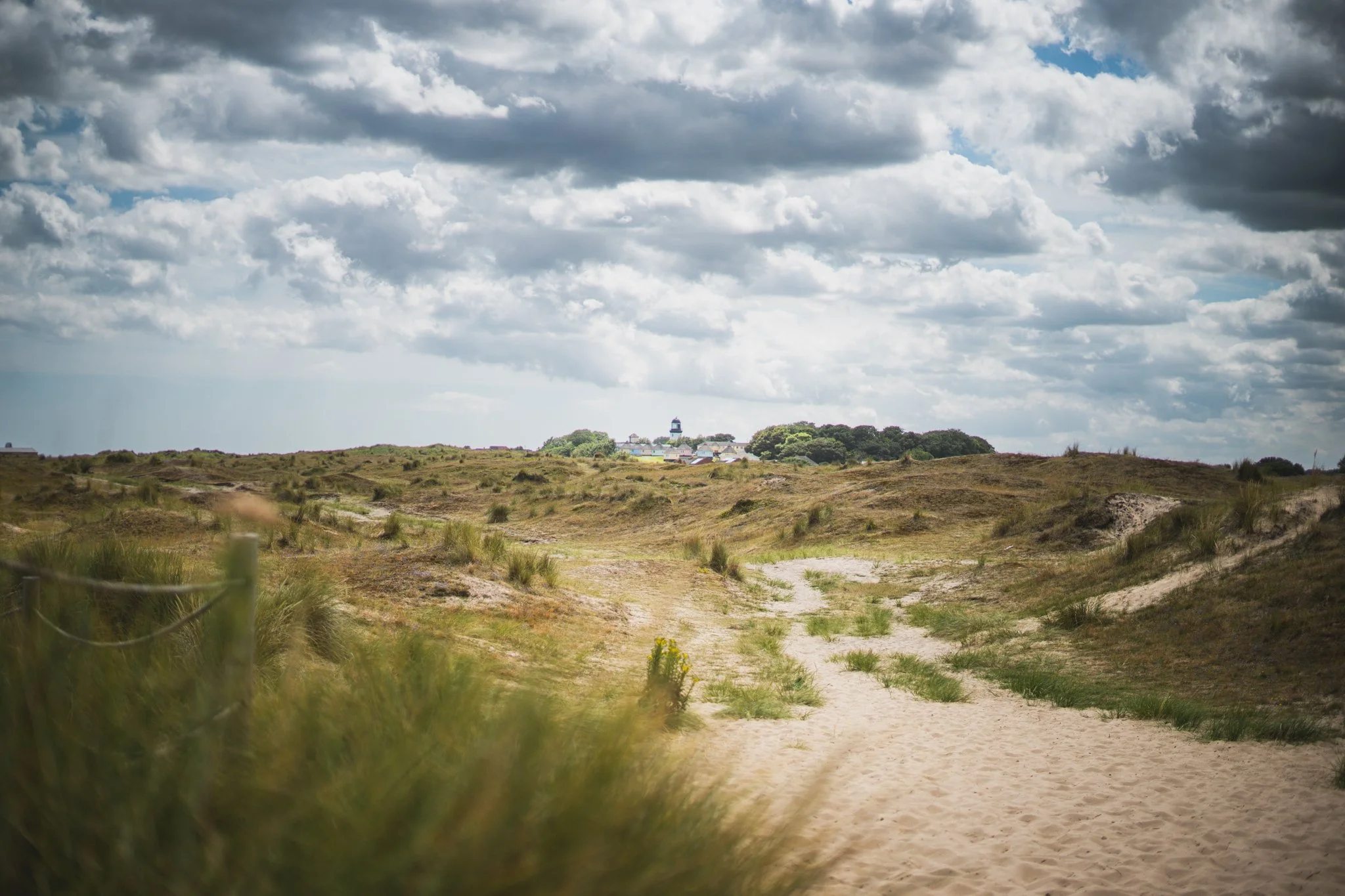 Sand dunes and grassy landscape under a cloudy sky near a small town or village with a lighthouse or tower in the distance.