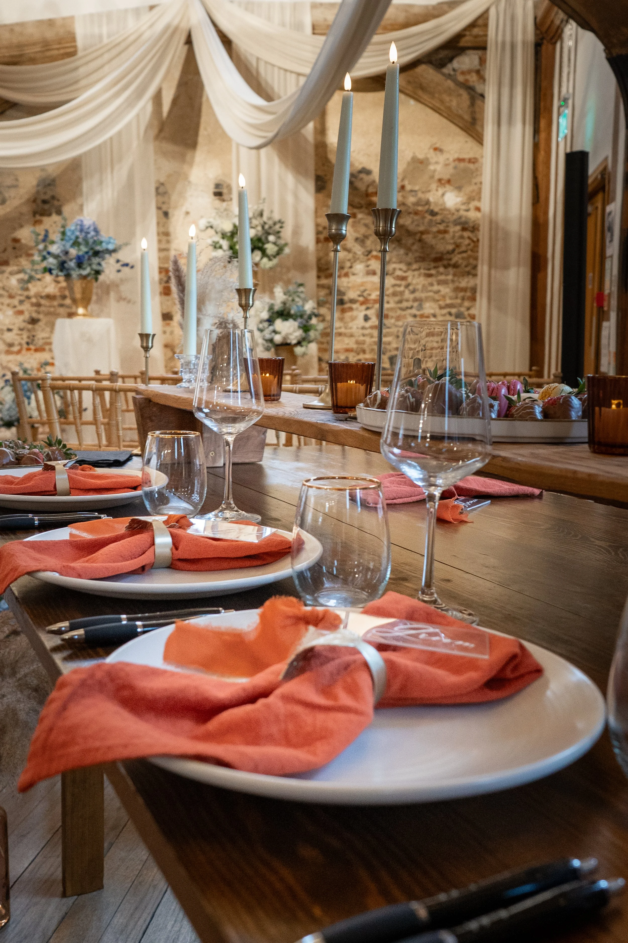 A decorated table set with orange napkins, wine glasses, candle holders with lit candles, and a platter of chocolates, in a rustic venue with exposed brick walls and floral arrangements.