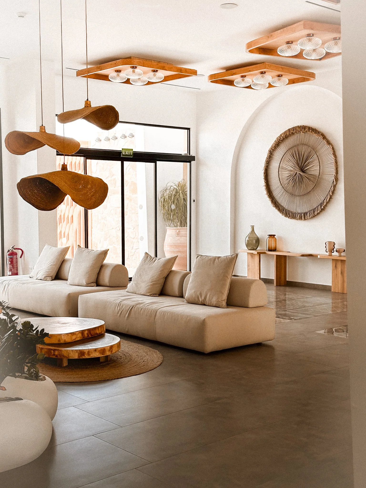 Modern living room with beige sofa, wooden coffee tables, wall art, and decorative vases. Mirrored ceiling lights and wooden pendant lights add to the decor. Large windows and a glass door allow natural light.