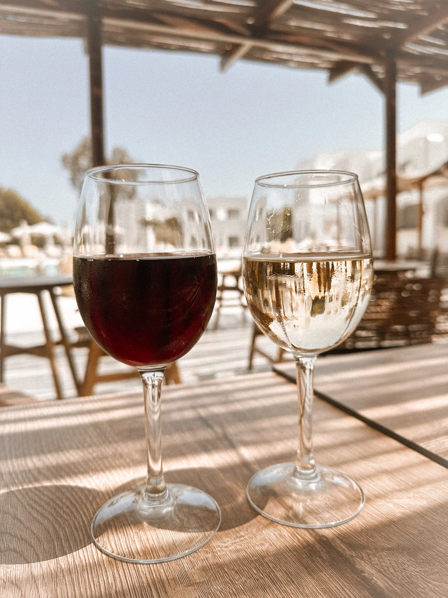 Two glasses of wine, one red and one white, on a wooden table outdoors with a blurred background of a patio with wooden furniture.