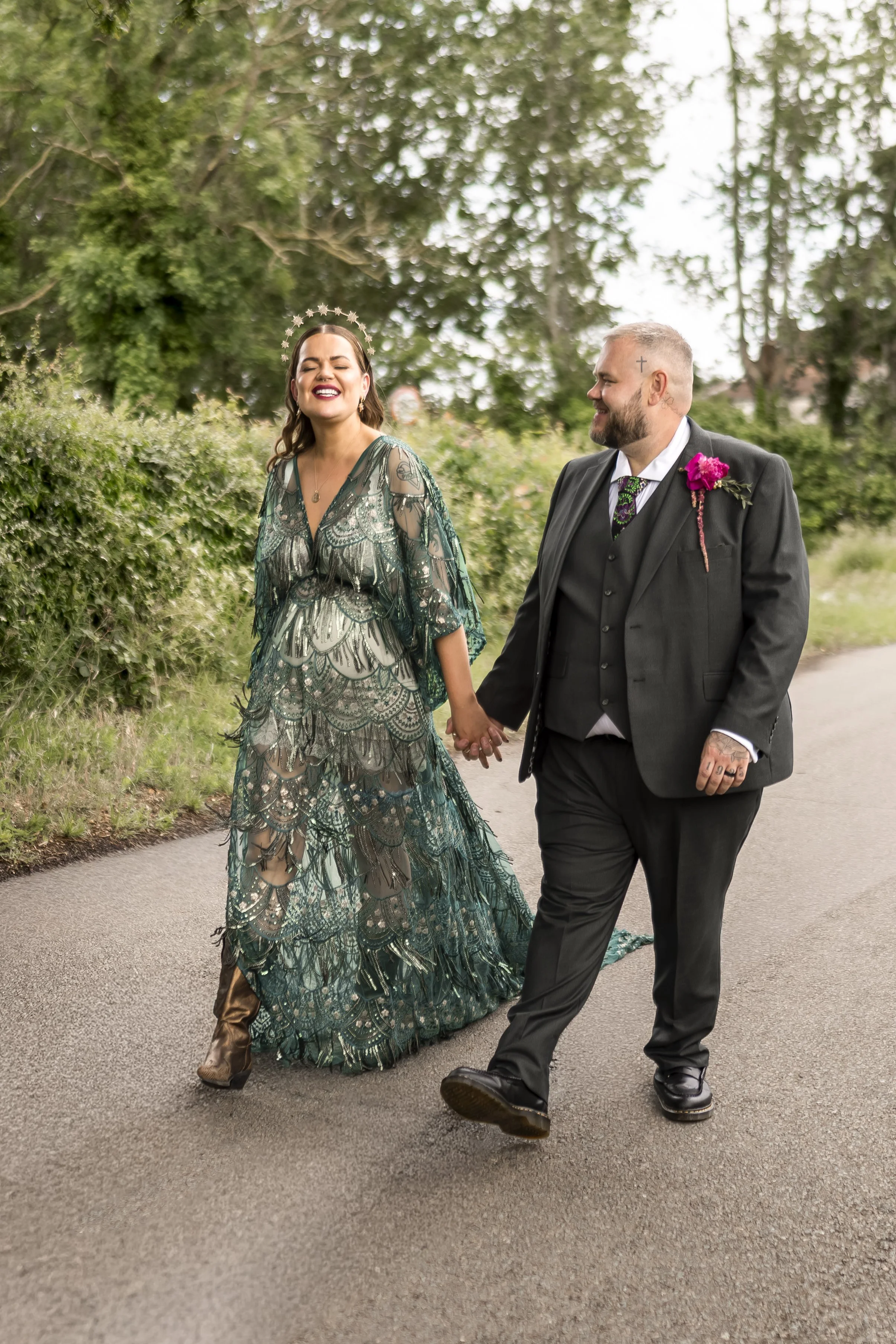A smiling woman in a detailed, green and blue patterned dress with a star-shaped headband, holding hands with a man in a black suit with a boutonnière, walking outdoors on a path with trees and greenery in the background.