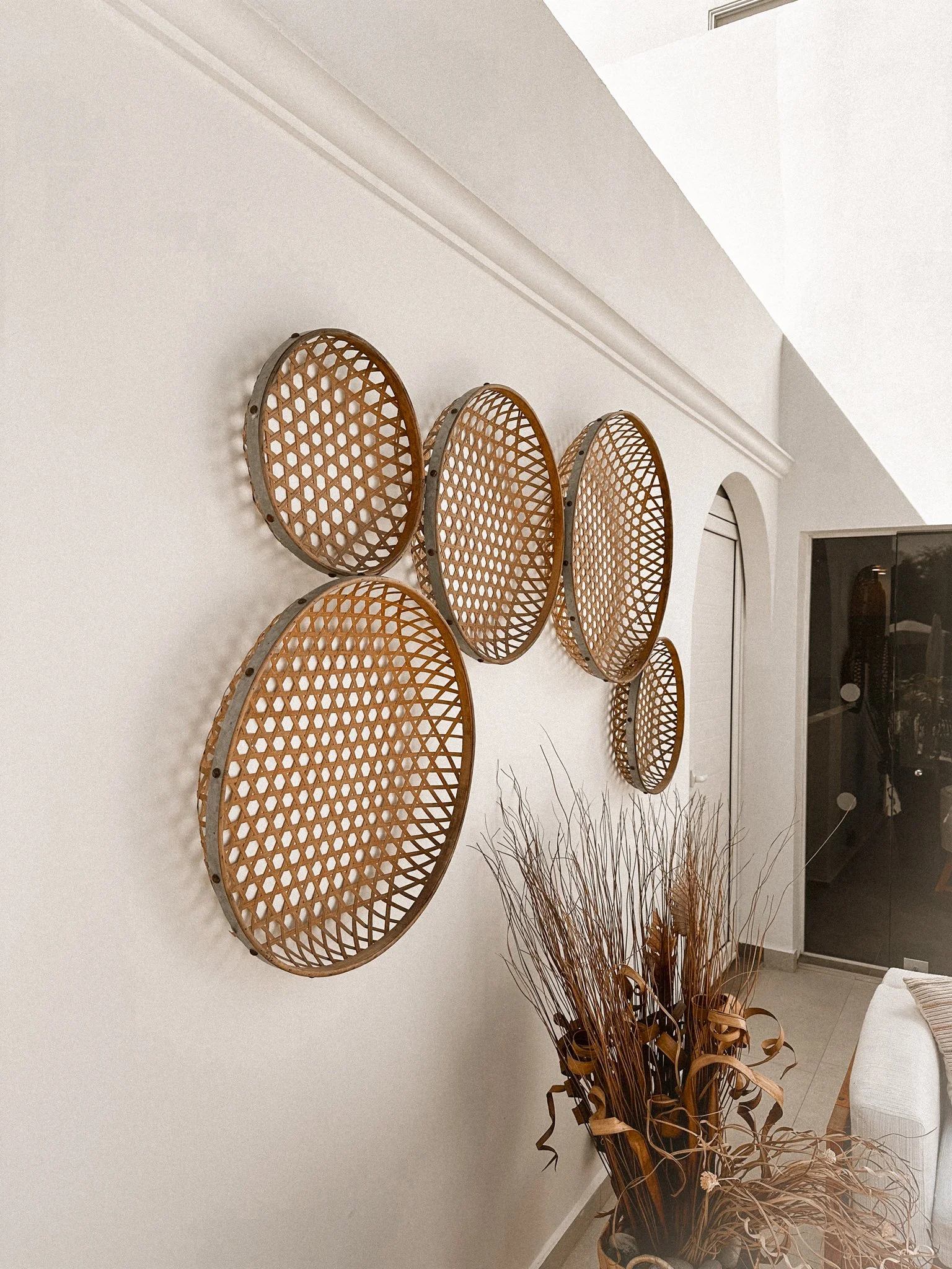 A decorative wall display featuring eight round, bamboo, lattice-like trays mounted on a white wall above a tall dried plant arrangement in a living room.