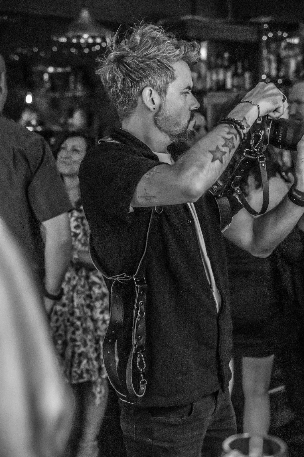A man with styled hair, tattoos, and a beard is taking a photo with a camera in a lively indoor setting, possibly a bar or a party, with blurred people in the background.