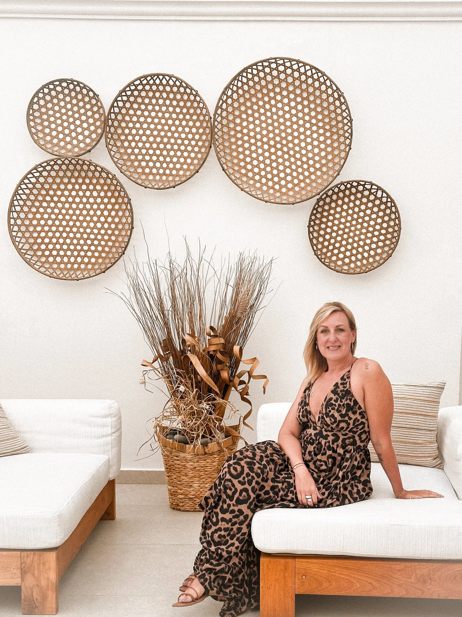 A woman with blonde hair and a leopard print dress sitting on a white cushioned bench in a modern, minimally decorated room. Behind her is a large basket with dried plants and a decorative wall display with six round wicker objects mounted on the wal