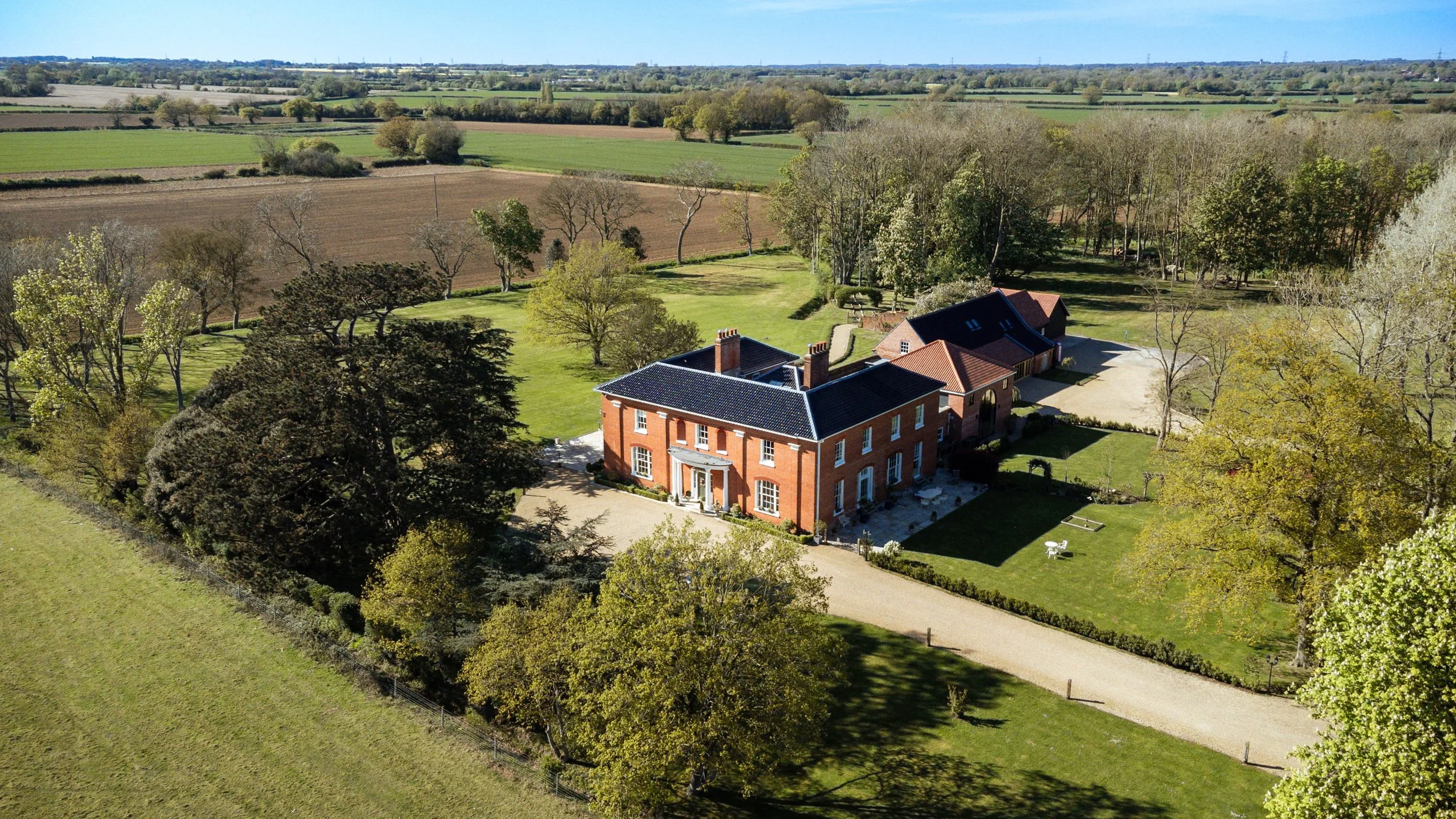 Aerial view of Reymonston hall with a dark roof, surrounded by trees and a well-maintained lawn, with expansive fields and open farmland in the background.