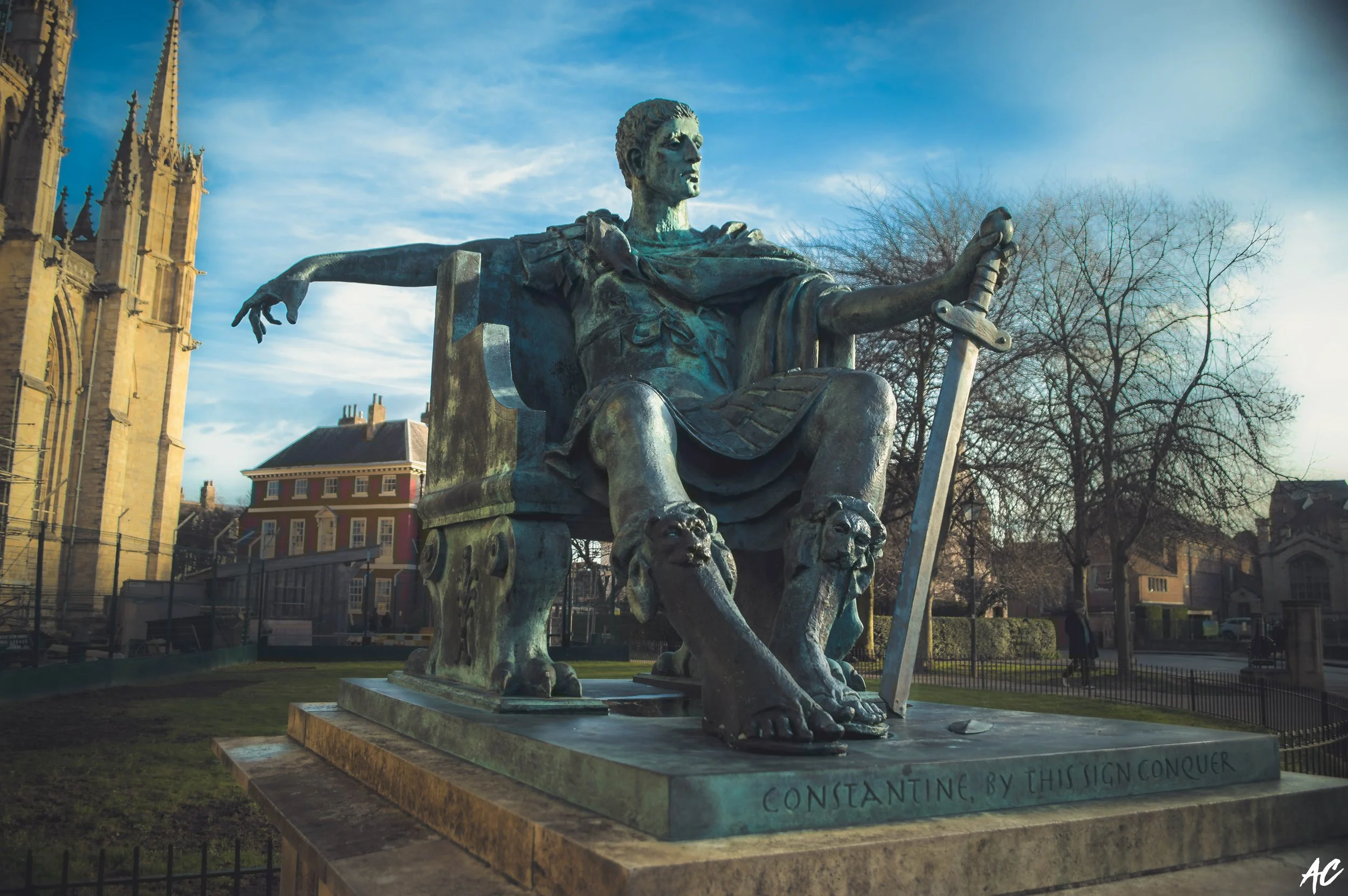 Bronze statue of Constantine the Great seated with a sword and a scroll, in a park area with historic buildings and leafless trees in the background.