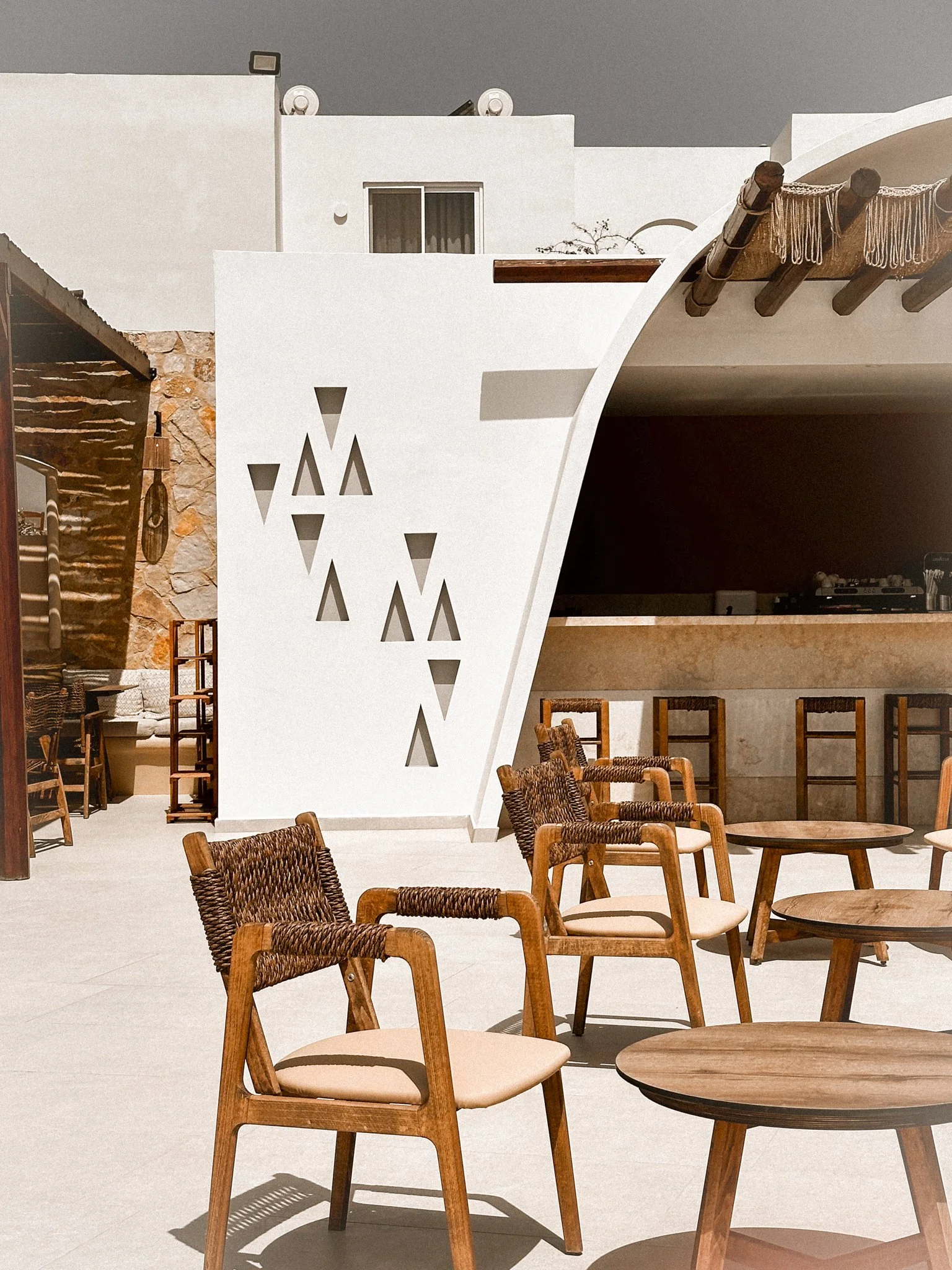 Outdoor seating area with wooden chairs and round tables, white modern building with geometric cutouts, stone and wood accents, and a shaded terrace with hanging ropes.