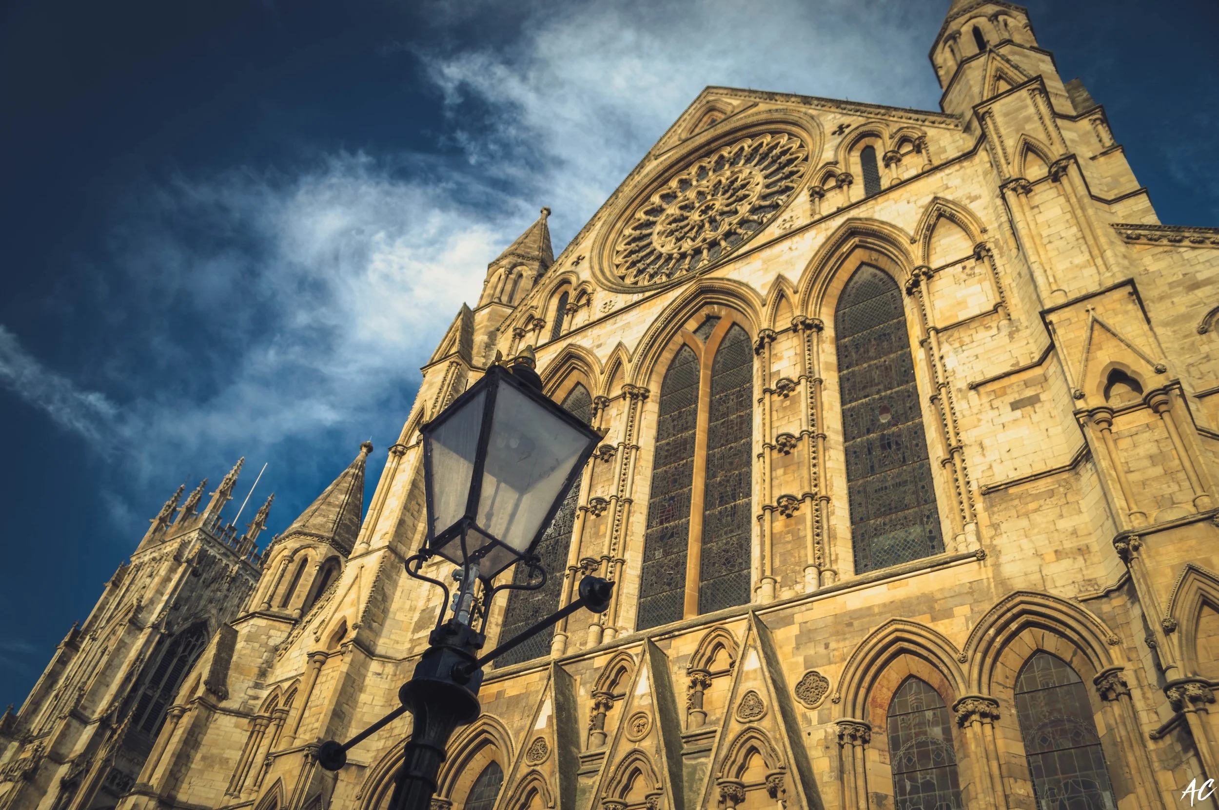 A historic Gothic cathedral with large stained glass windows and ornate stonework, seen from below against a partly cloudy sky, with a street lamp in the foreground.