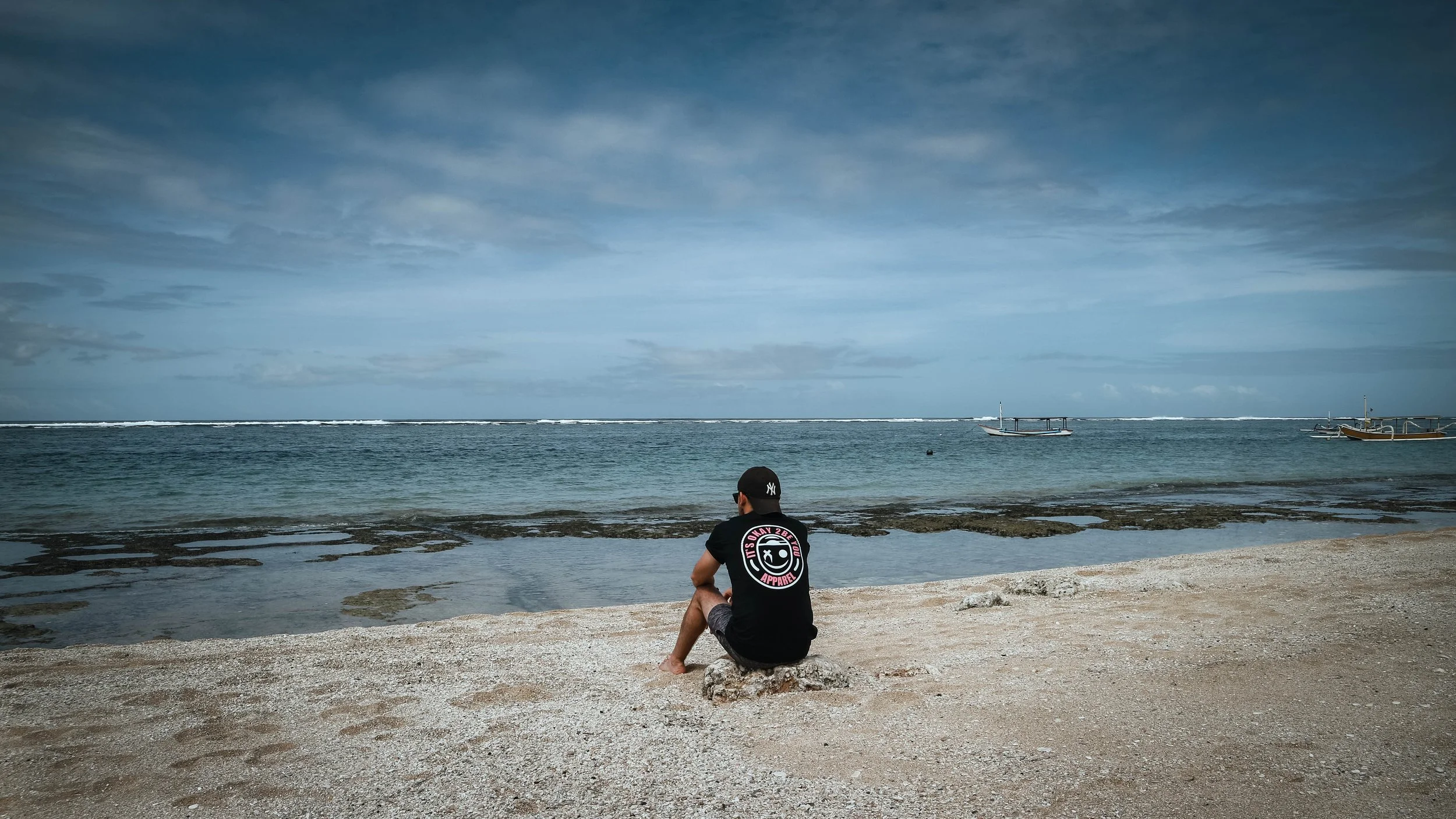 A person sitting on a rock at the beach, looking at the ocean with boats in the water and a cloudy sky above.